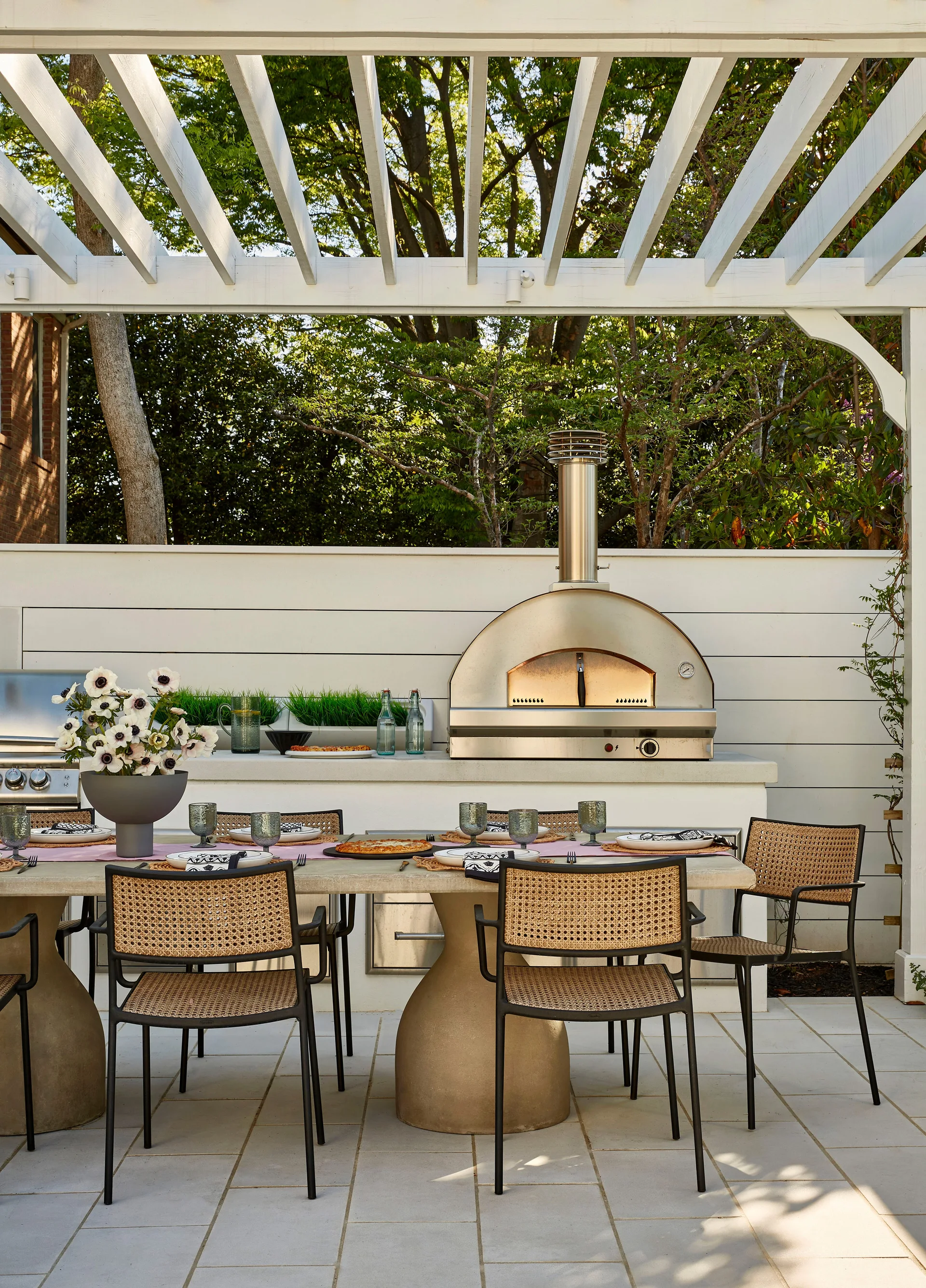 An outdoor dining patio features a concrete table with cane chairs under a white pergola next to a stainless steel pizza oven.