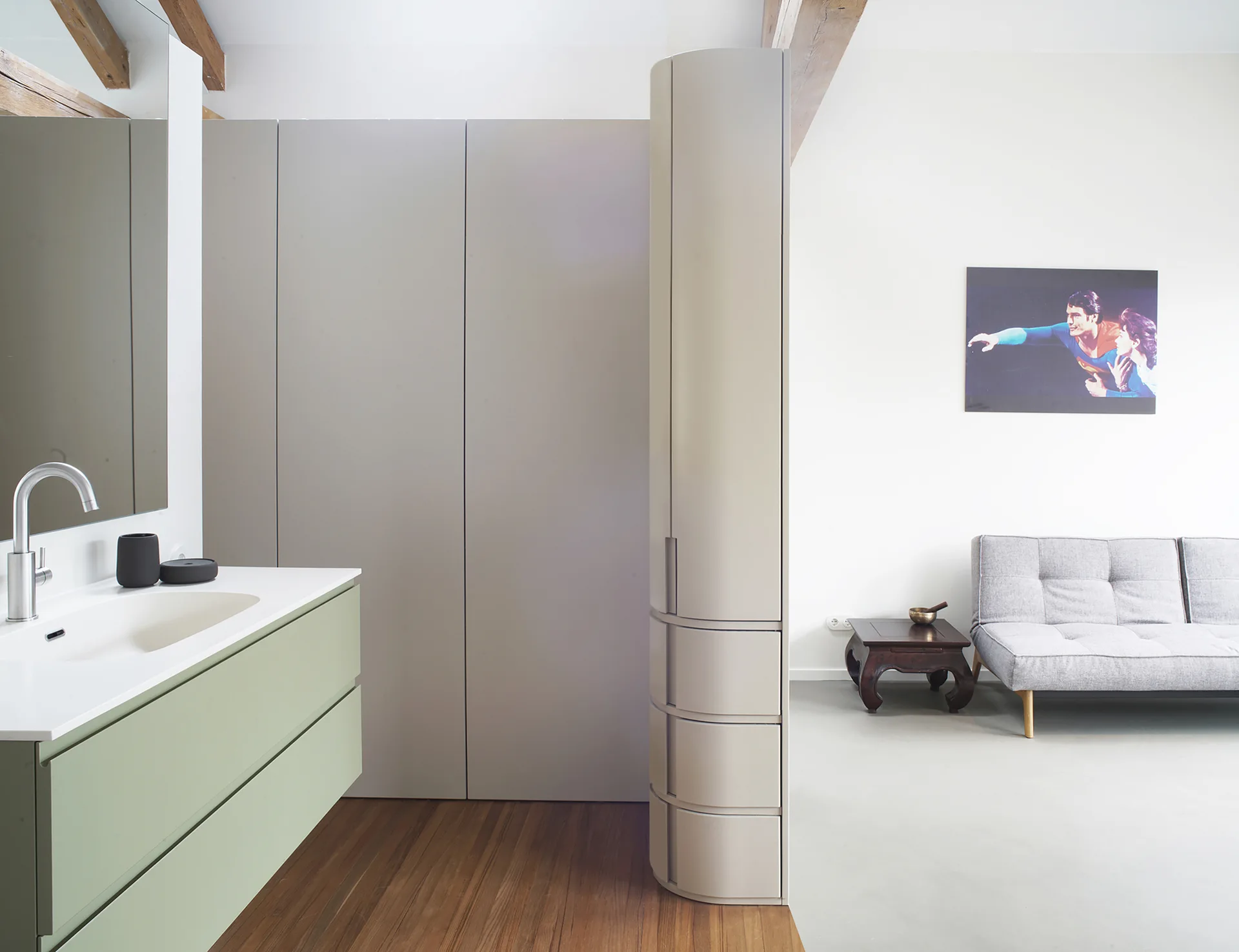 A sage green bathroom vanity with a white sink transitions to a living space featuring a tall curved storage tower, wood flooring, and a grey sofa.