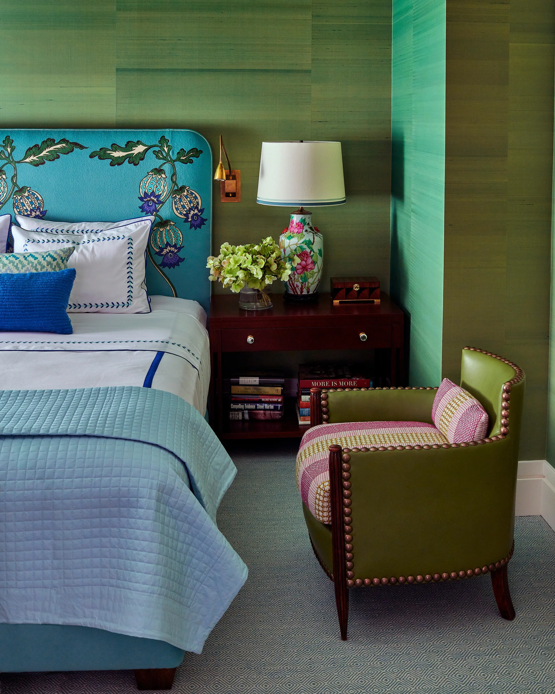 A bedroom features green grasscloth walls, a teal floral headboard, a dark wood nightstand with a lamp, and a green leather armchair with nailhead trim.