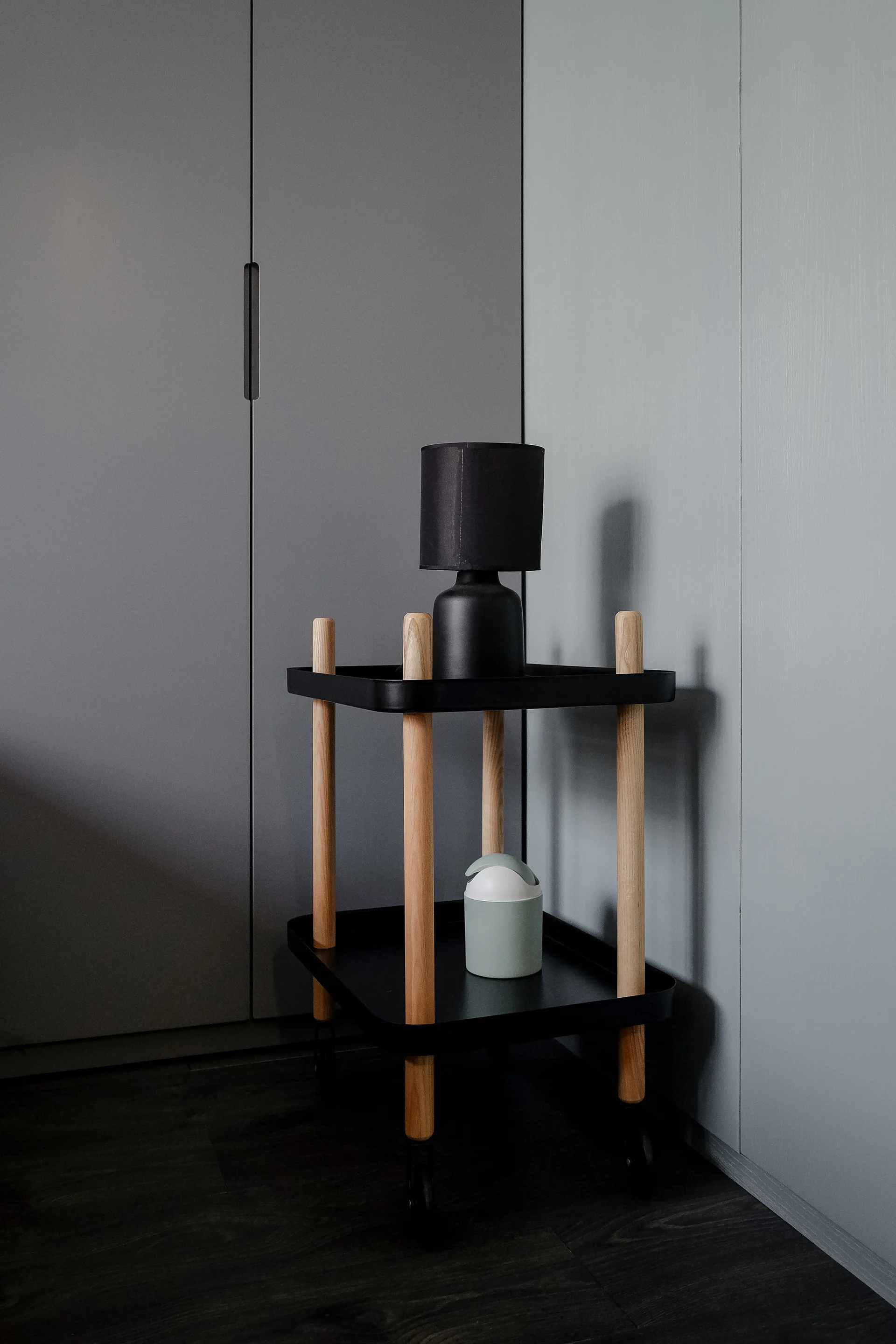 A two-tier black tray cart with light wood legs stands in front of grey closet doors, holding a black lamp and a small sage green container.