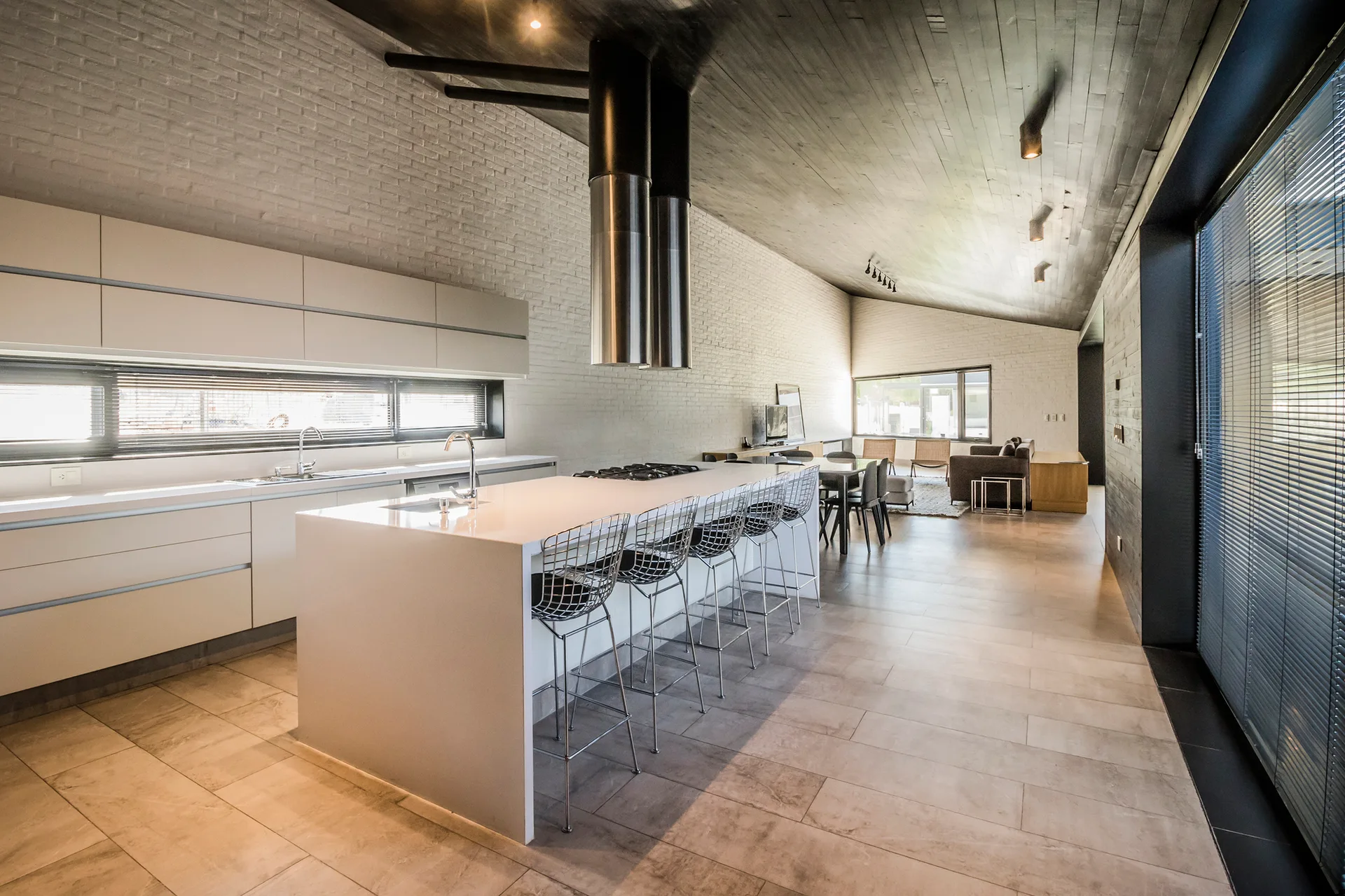 An open-plan kitchen features a large white island with wire bar stools, white brick walls, and a dark slanted wood ceiling.