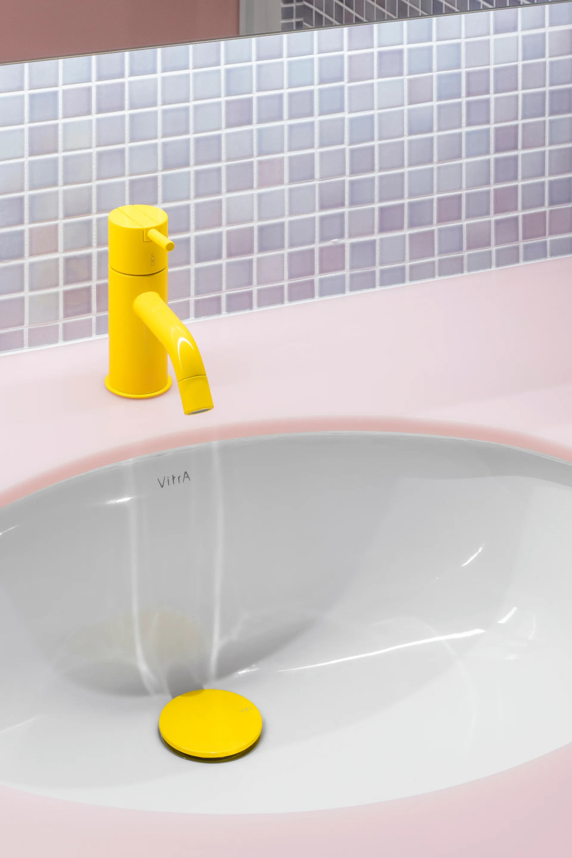 A close-up of a pink bathroom countertop featuring a bright yellow faucet, matching drain plug, and lavender mosaic tile backsplash.