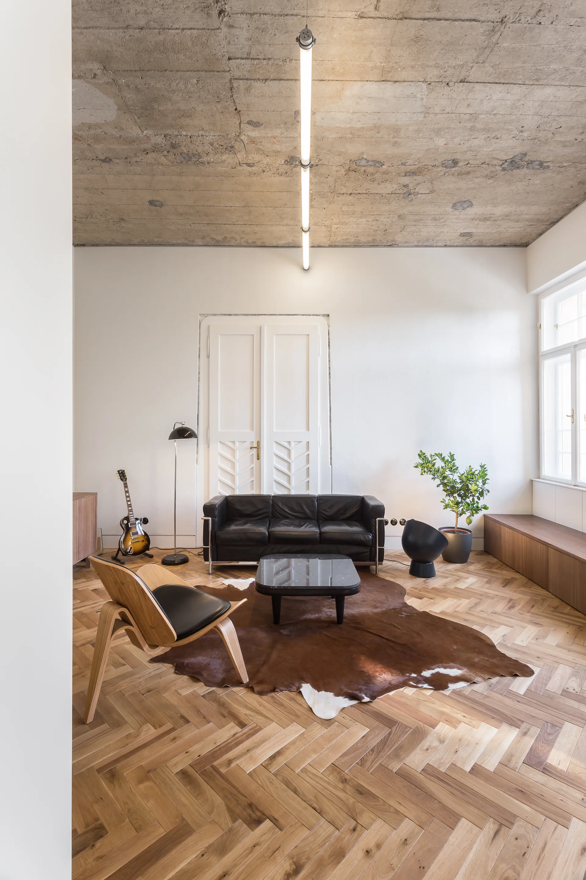 A minimalist living room with a black leather sofa and wood armchair on a herringbone parquet floor beneath an exposed concrete ceiling.