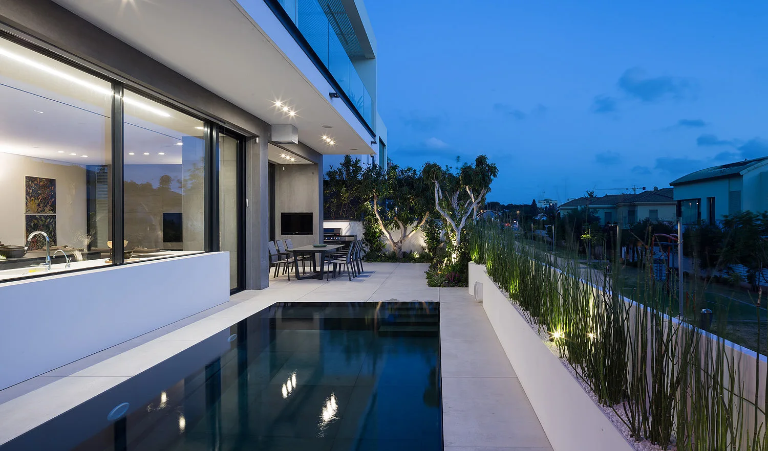A modern backyard patio features a narrow rectangular swimming pool alongside a white planter with tall grasses illuminated by landscape lighting at twilight.