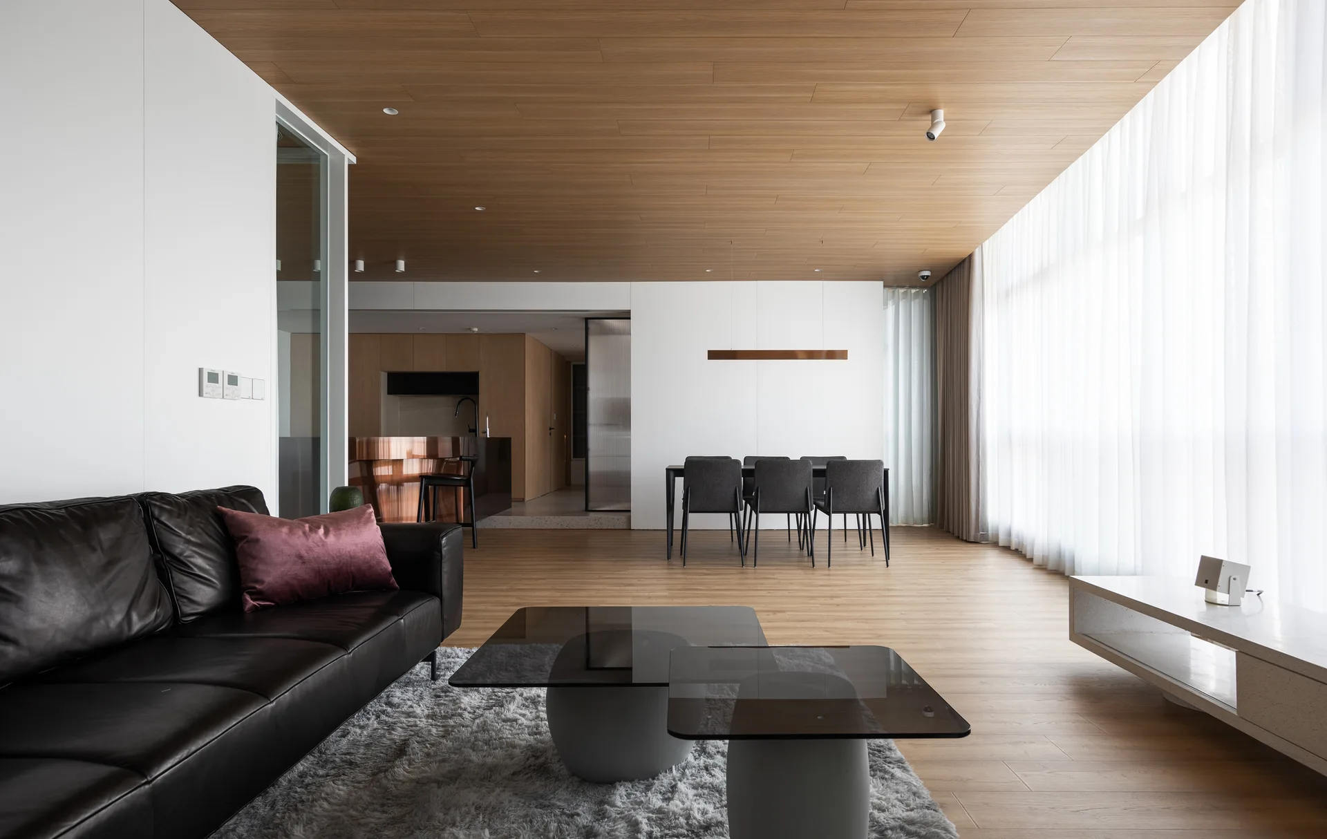 An open-concept living room features a wood paneled ceiling, a black leather sofa on a shag rug, and a dining set near sheer curtains.