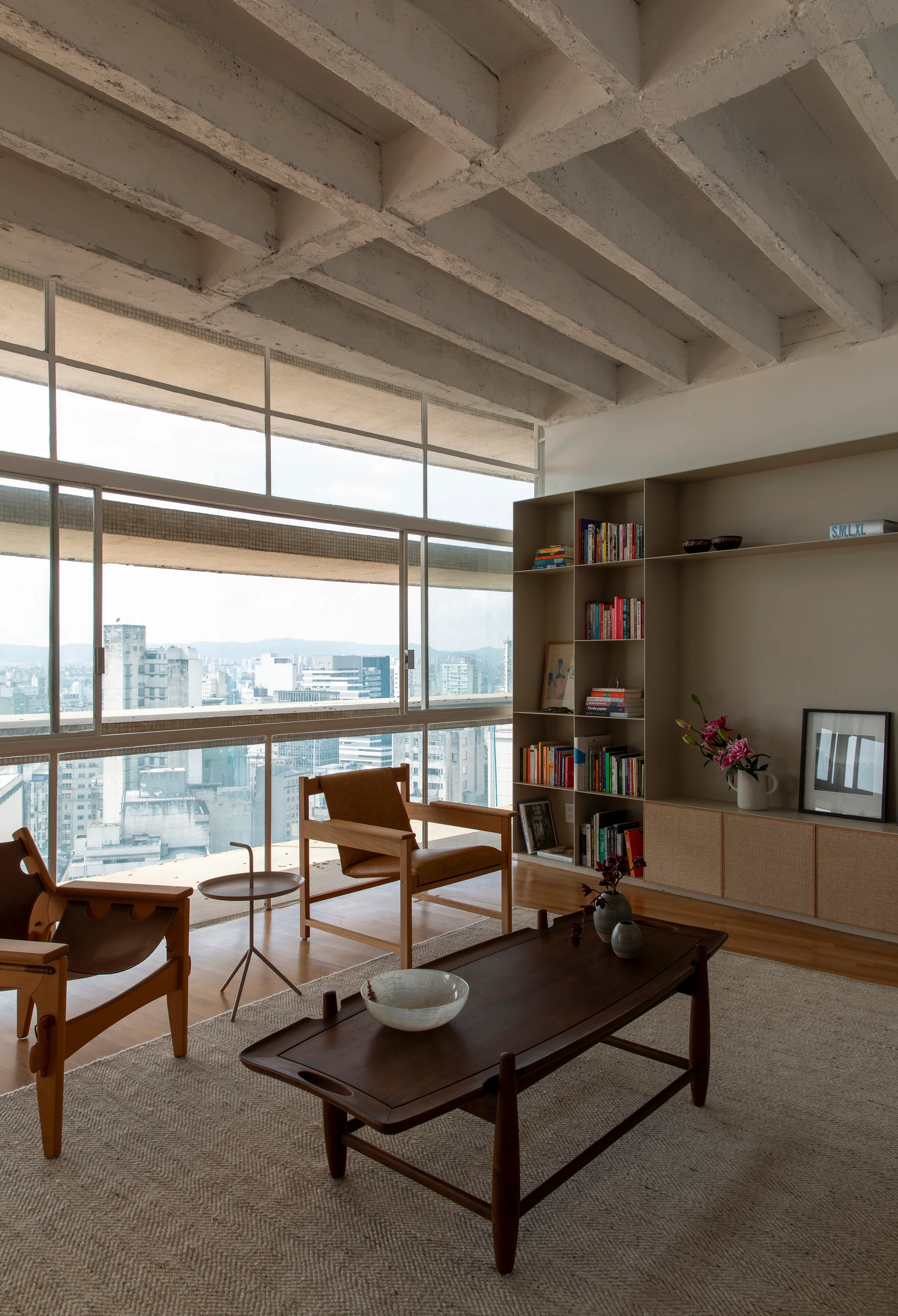 A modern living room features wood armchairs, a low coffee table, beige built-in shelving, and an exposed concrete waffle ceiling with large windows.