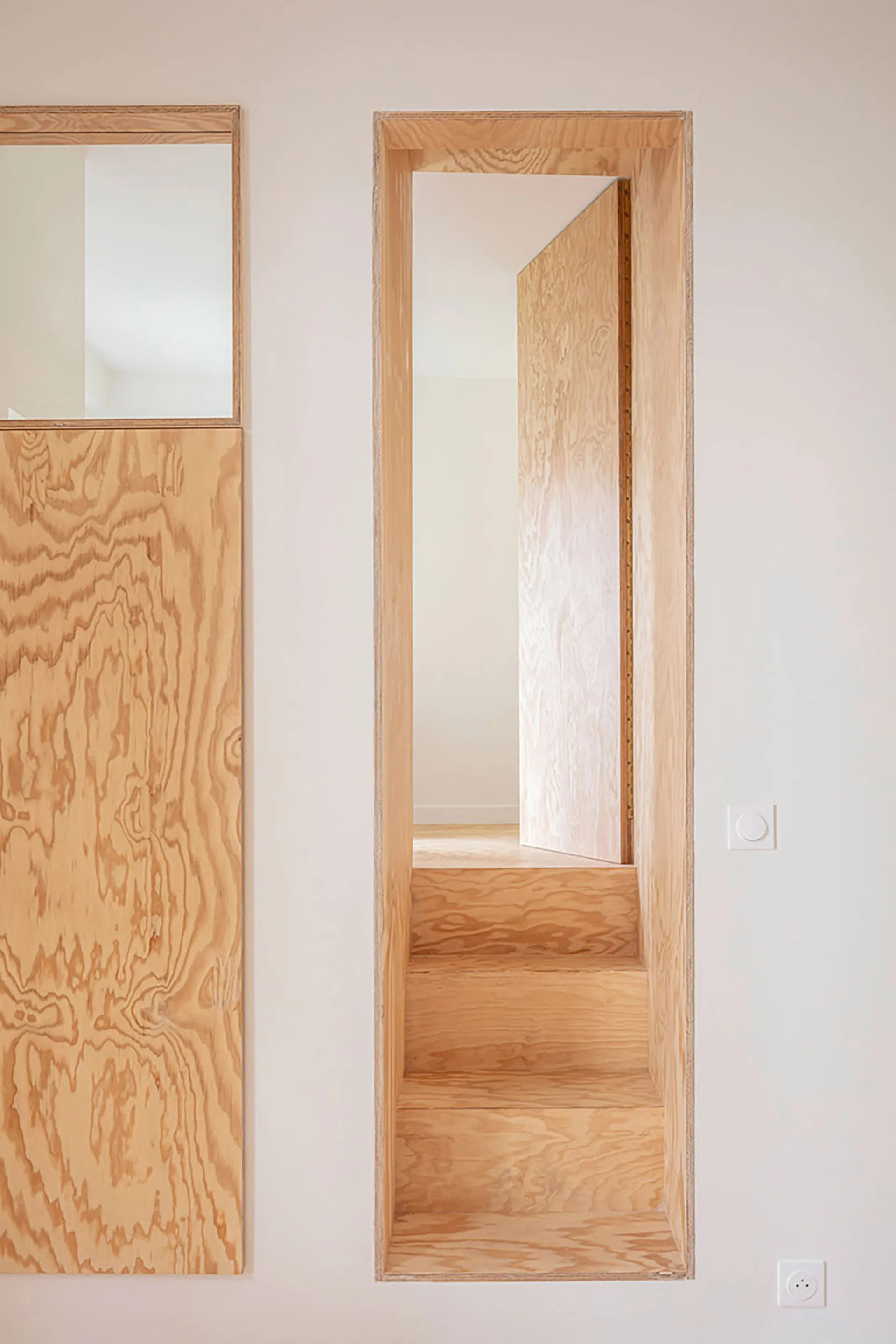 A narrow plywood staircase is built into a tall wooden door frame on a plain white wall next to a matching wood panel.