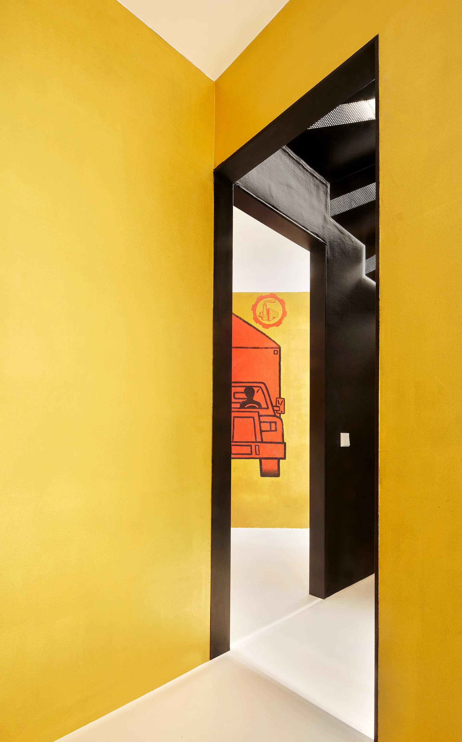 A bright yellow hallway features a wide matte black door frame looking into a room with white floors, a black staircase, and an orange truck mural.