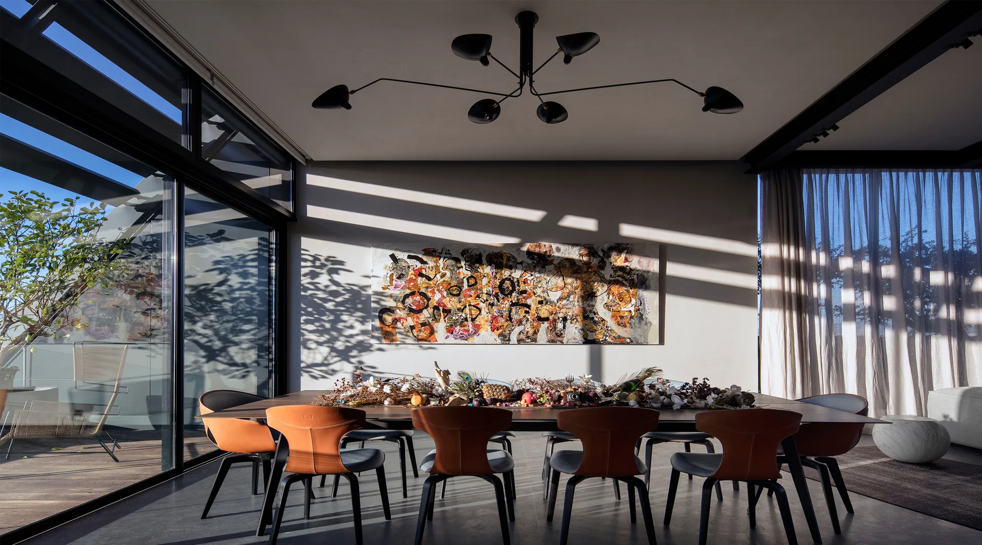 A modern dining room features a long table surrounded by tan leather chairs under a multi-arm black chandelier, with a large abstract painting on the wall.