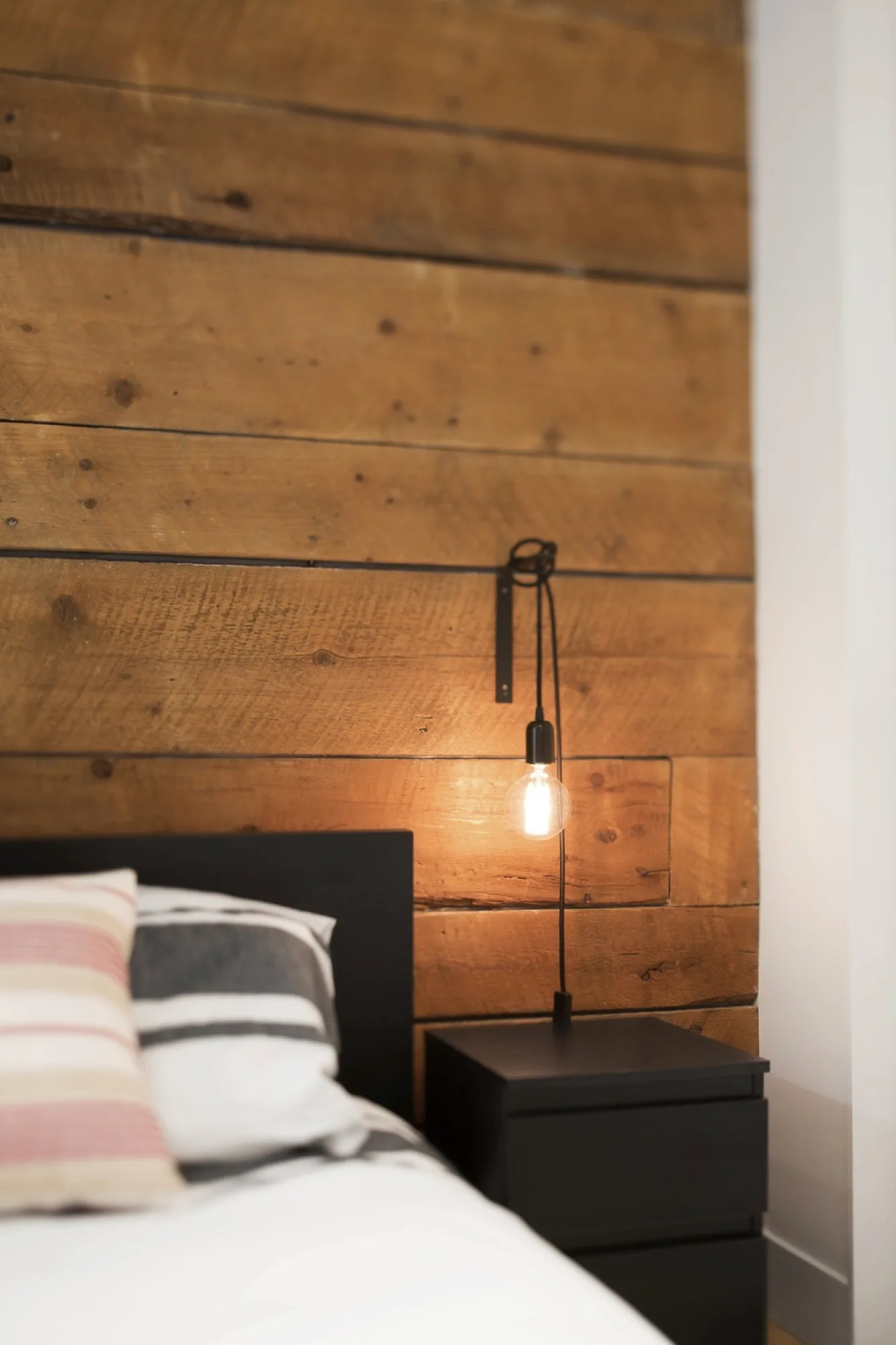 A black headboard and matching nightstand stand against a textured wood-plank accent wall with a hanging exposed-bulb light fixture.