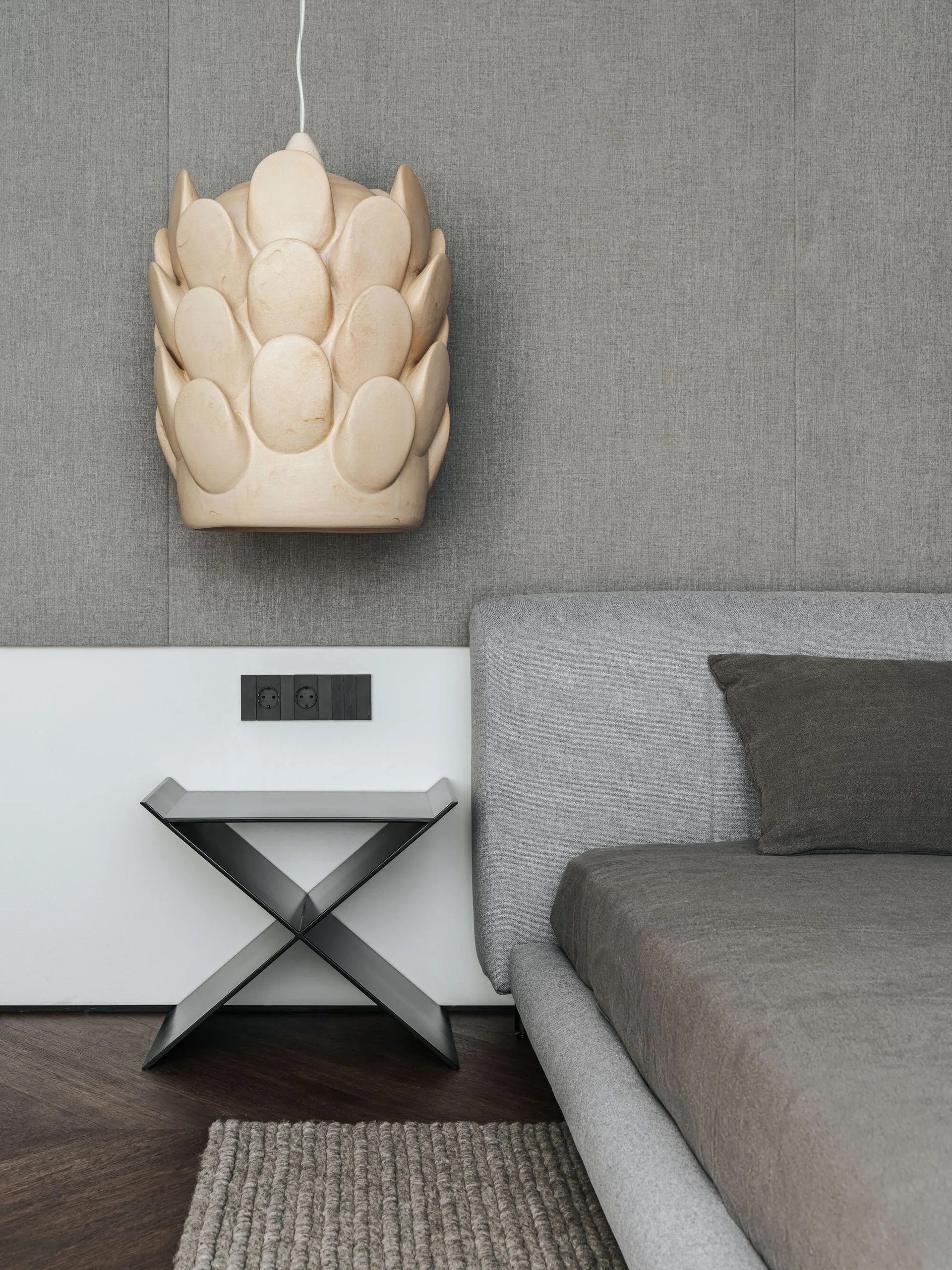 A grey upholstered bed sits beside a black x-frame side table beneath a textured, pinecone-shaped pendant light and grey fabric wall paneling.