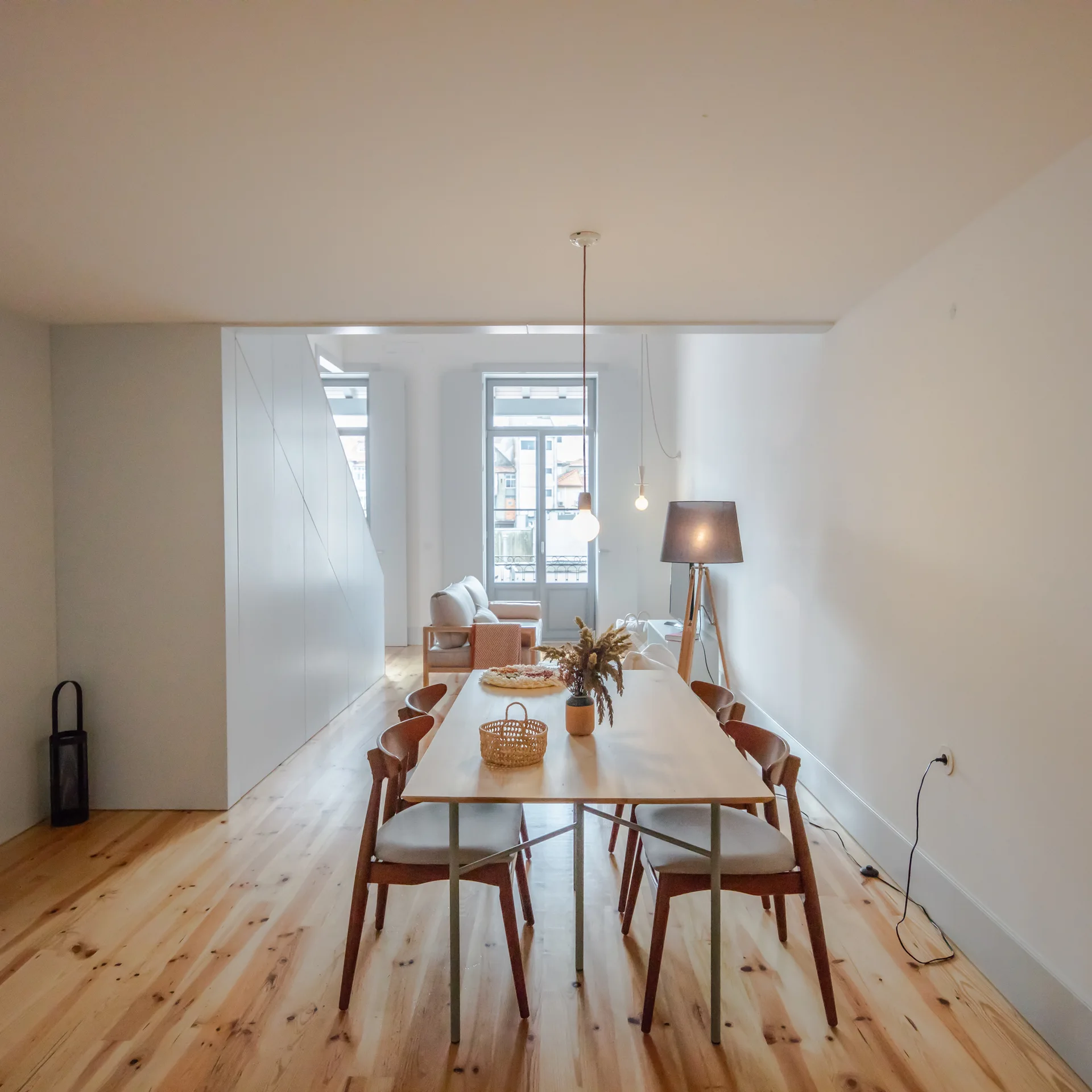 A minimalist dining area features a light wood table, mid-century chairs, natural wood floors, and a white slanted built-in storage wall.