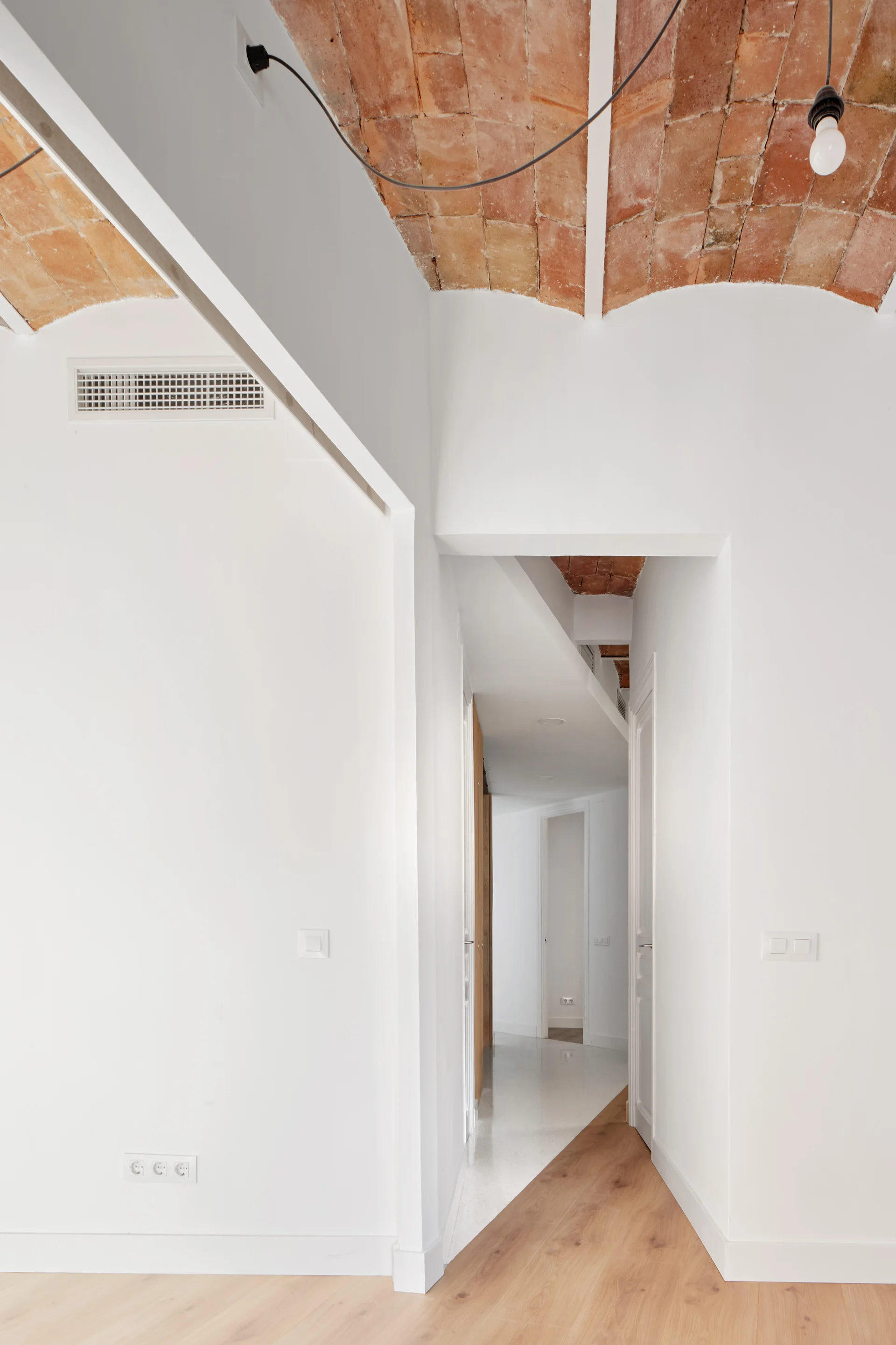A bright white hallway features a vaulted terracotta brick ceiling, light wood flooring, and minimalist white walls leading to adjacent rooms.