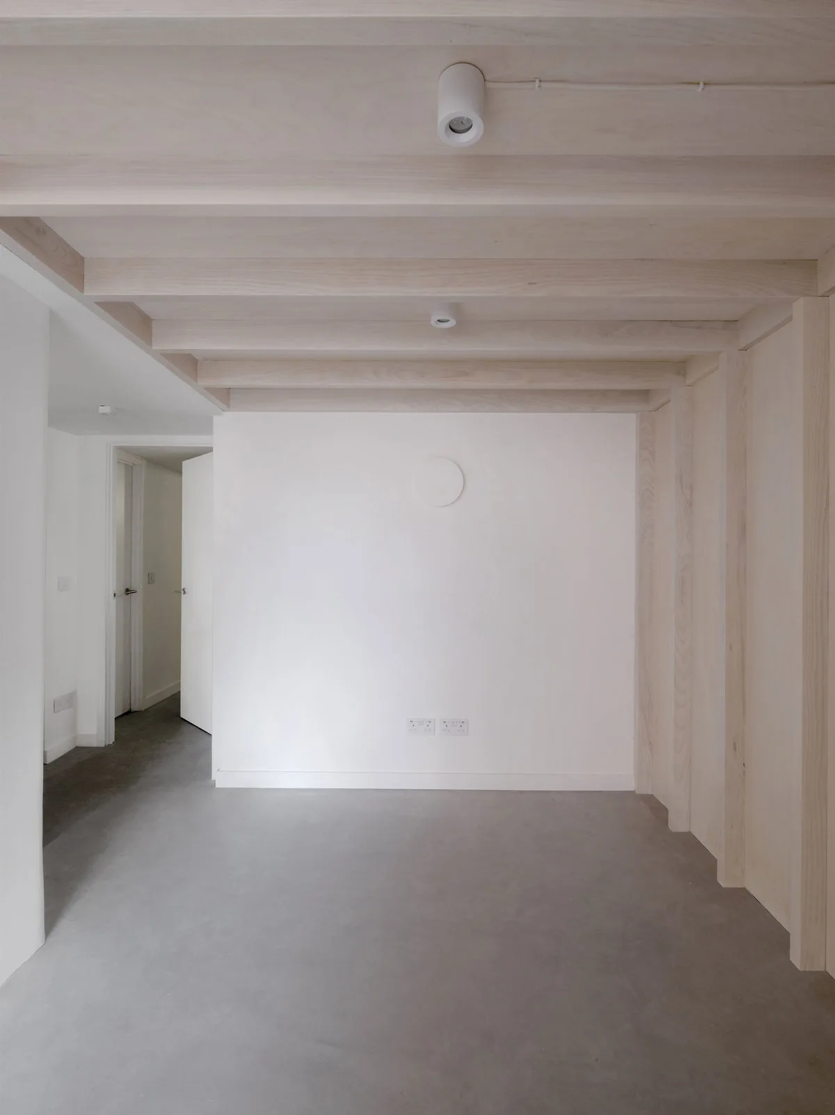 A minimalist room with exposed light wood ceiling beams, smooth concrete flooring, white plaster walls, and cylindrical ceiling spotlights.