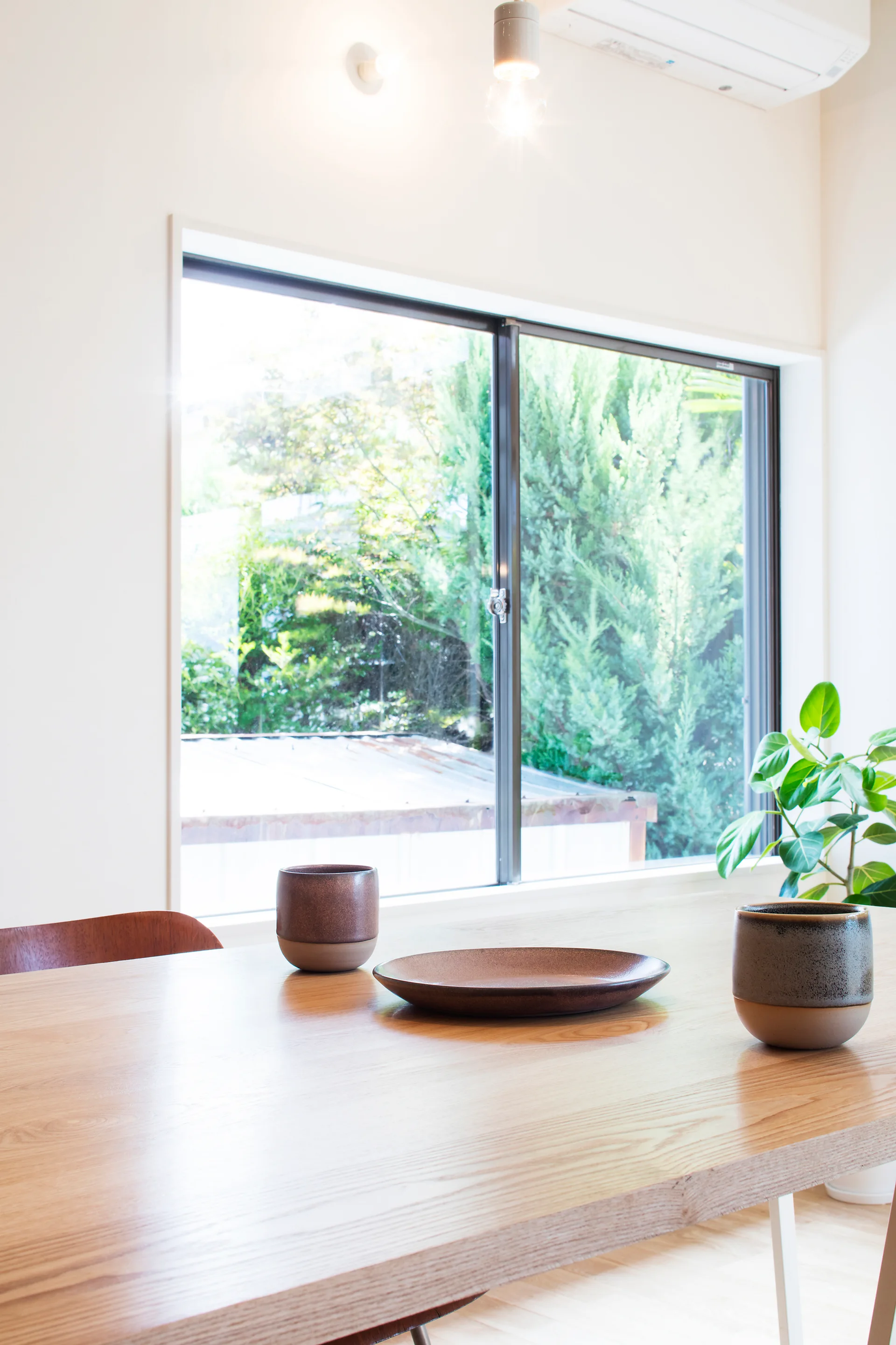 A light wood dining table with two textured brown ceramic cups and a plate sits in front of a window overlooking green garden foliage.