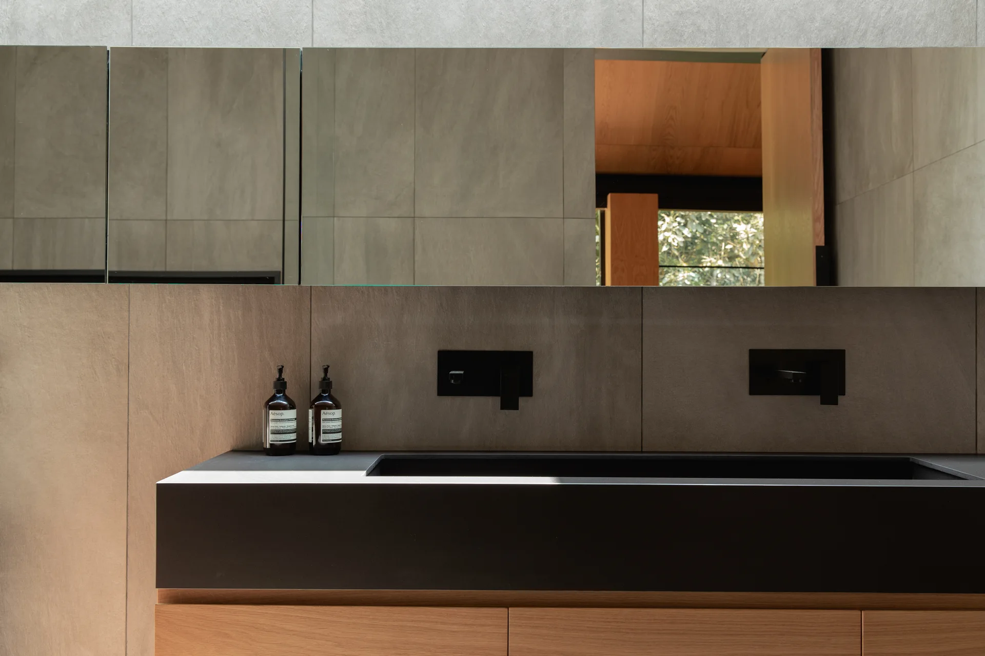 A modern bathroom vanity features a warm wood cabinet base, a matte black integrated sink, wall-mounted black faucets, and large grey tiled walls.