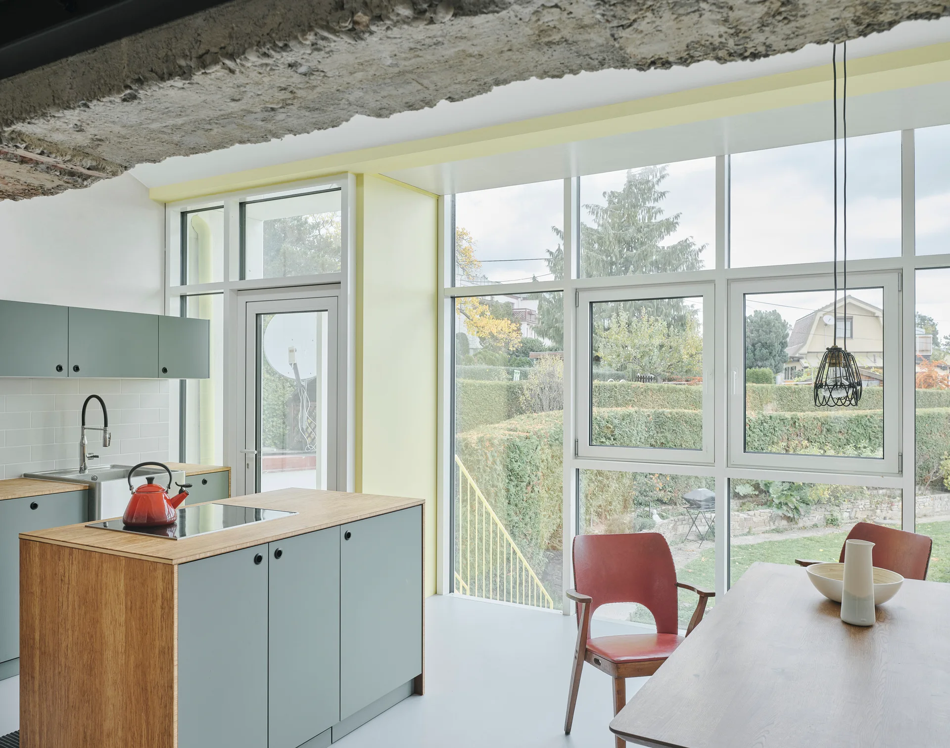 Sage green kitchen cabinets and a wood-topped island sit beneath an exposed concrete beam next to large windows overlooking a green garden.