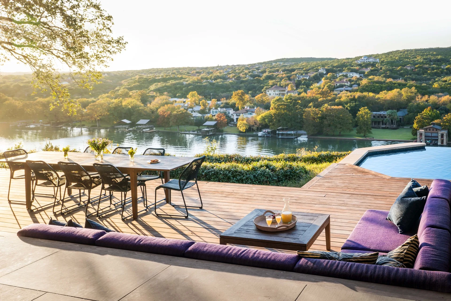 A spacious wood deck features a large purple sectional sofa, a wooden outdoor dining set, and an infinity pool overlooking a scenic lake landscape.