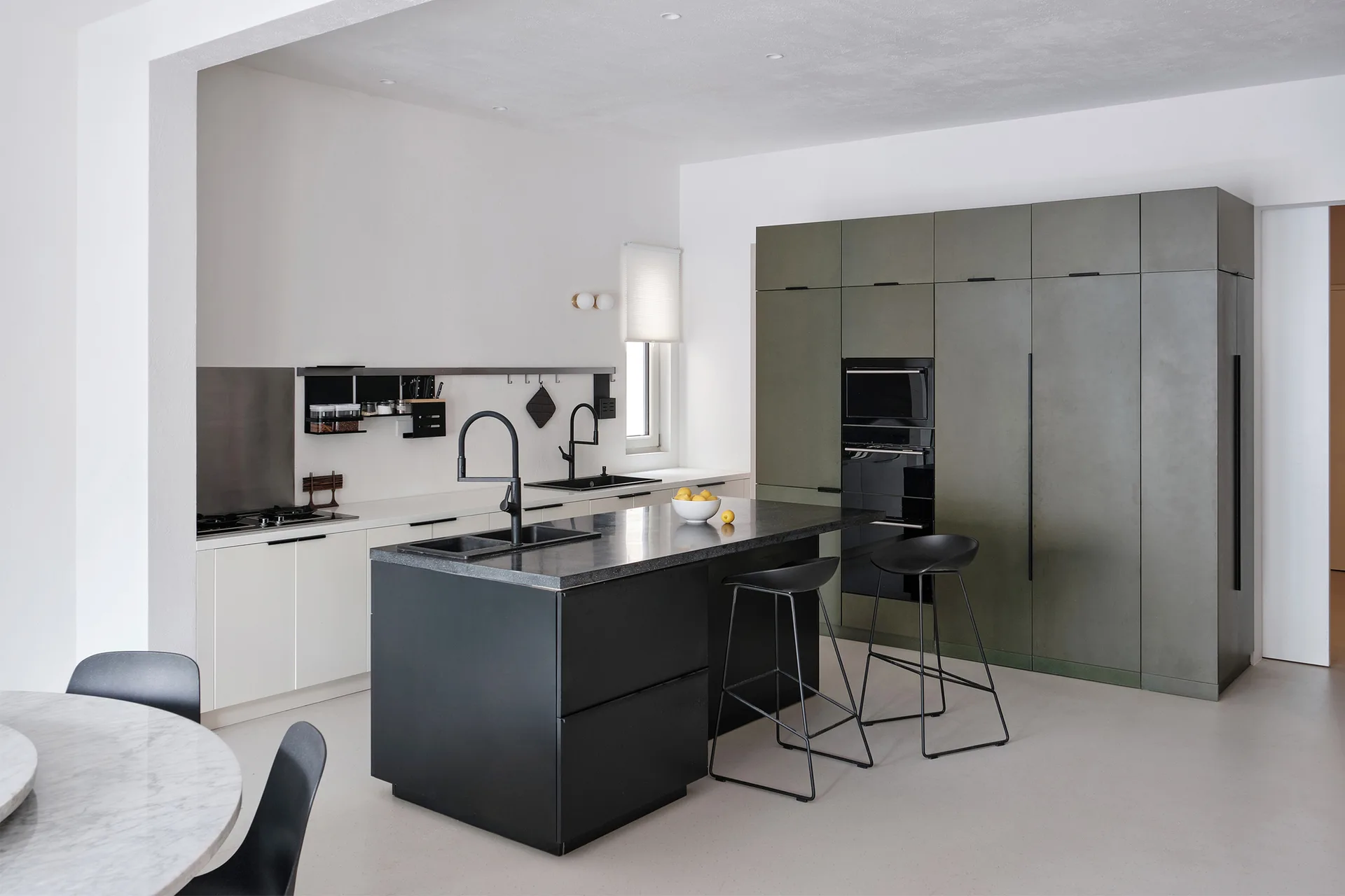 A minimalist kitchen features a matte black central island with stools, flat-front olive green cabinetry, and white walls.