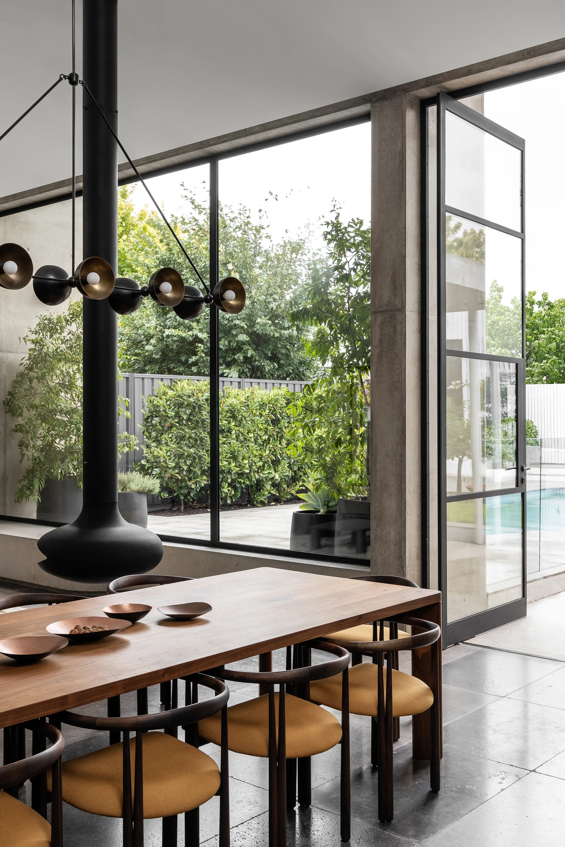 A wooden dining table with mustard yellow chairs sits near a suspended black fireplace and large glass pivot door opening to an outdoor pool area.