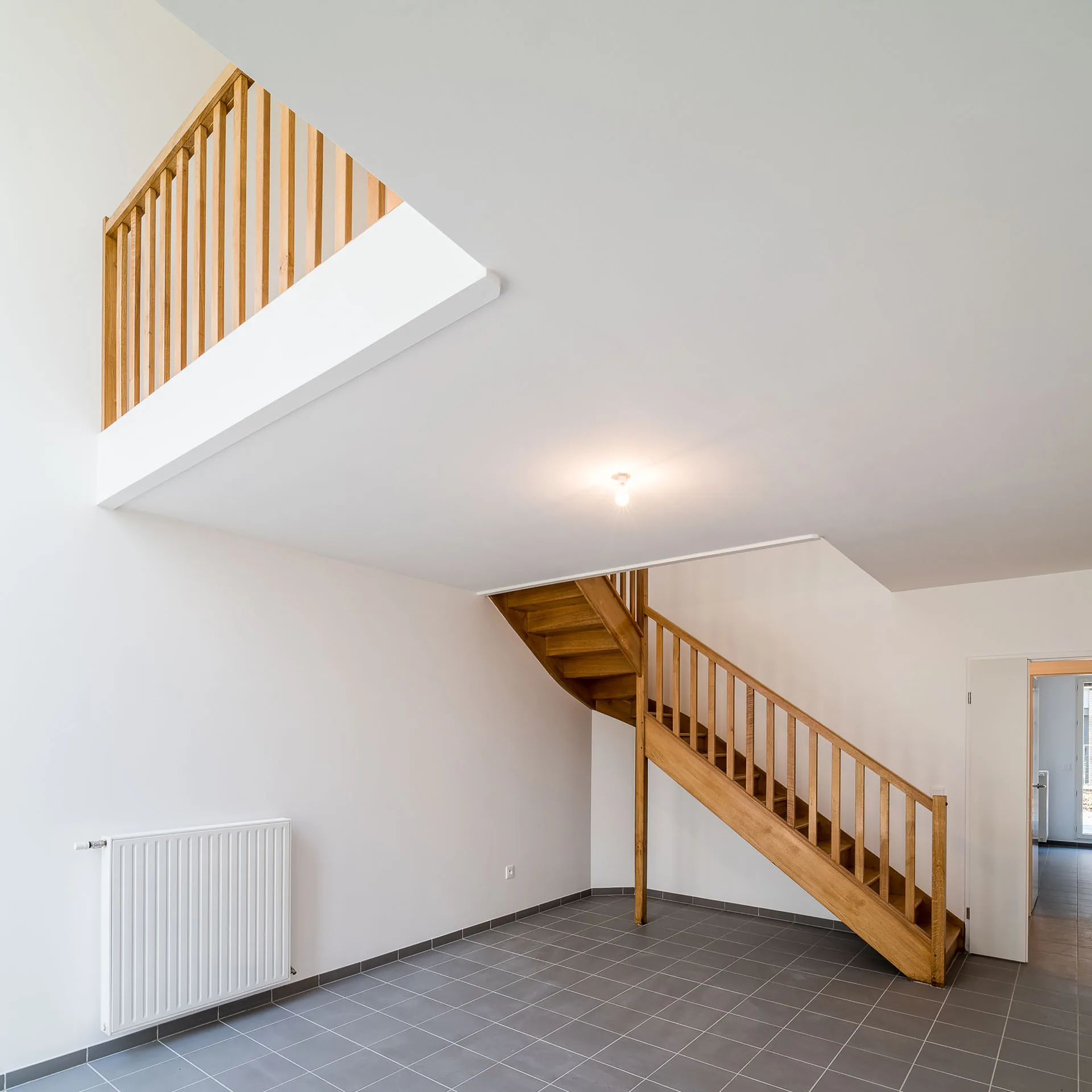A wooden staircase with a balustrade leads up to a mezzanine level in an empty room with white walls and grey tiled floors.