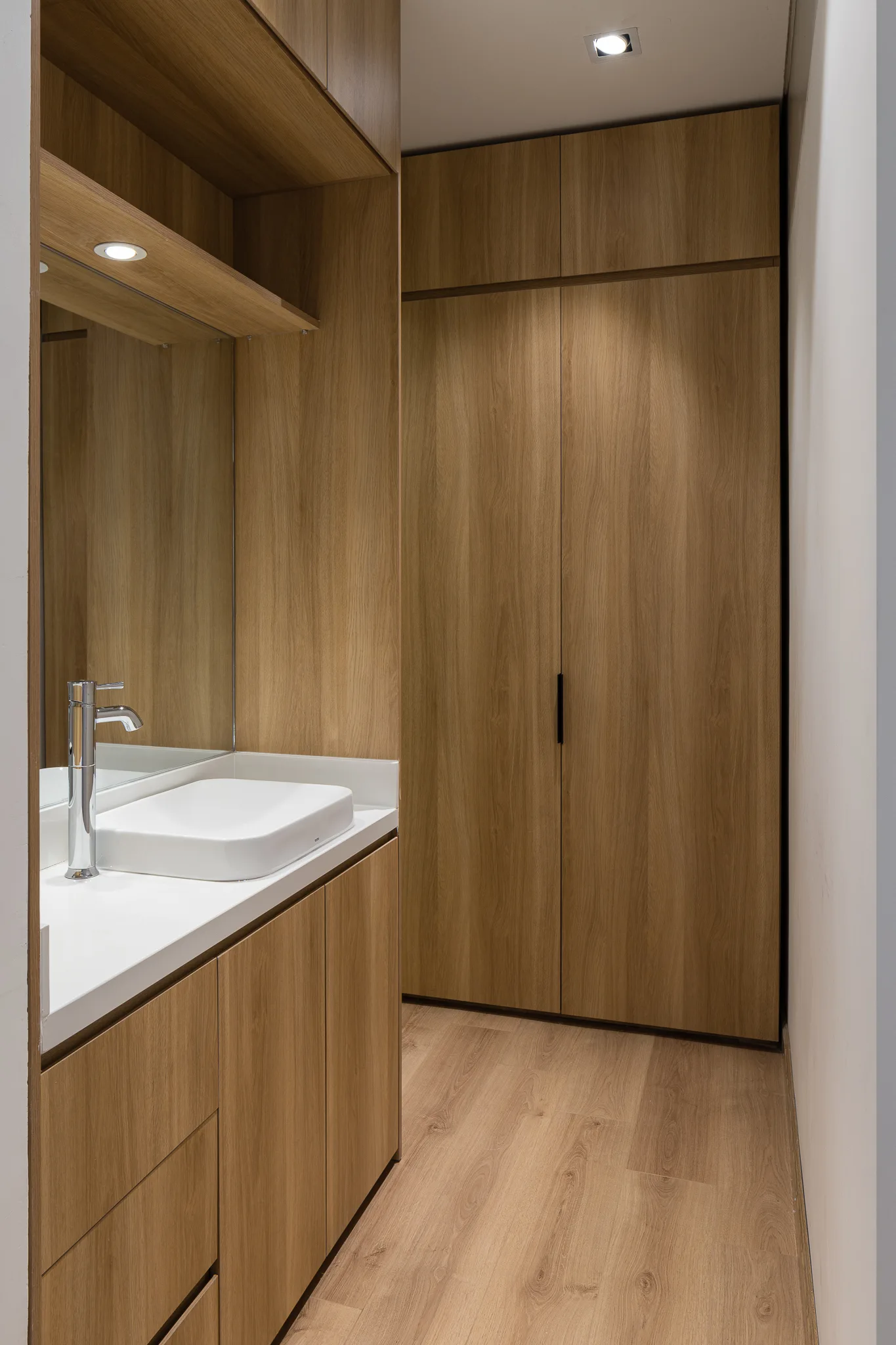 A modern bathroom featuring built-in wood grain closets, a matching wood vanity with a white countertop, chrome faucet, and wood flooring.
