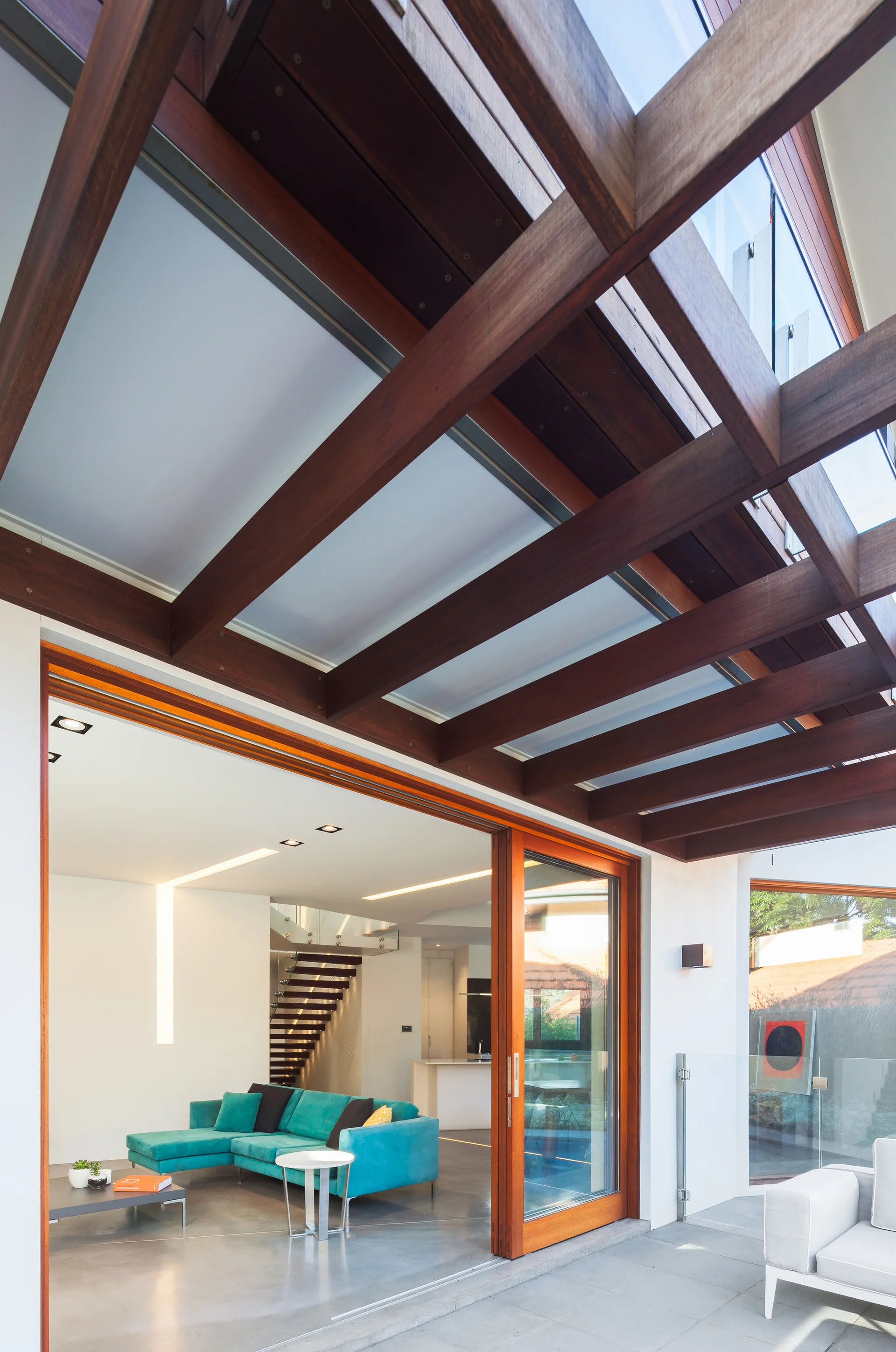 A modern living room with a teal sectional sofa opens onto a patio beneath a dark wood pergola through large sliding glass doors.