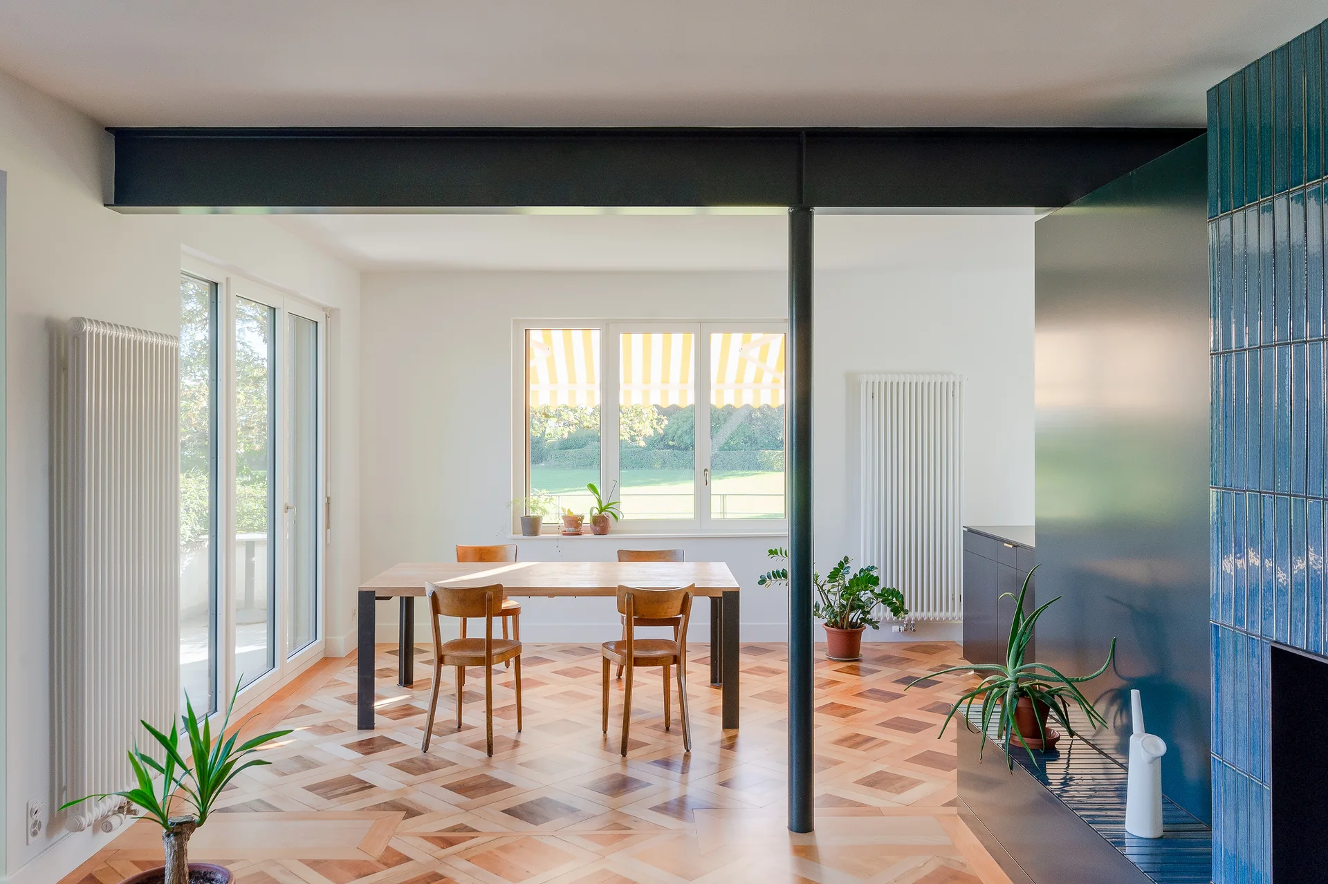 A bright dining room features a wooden table and chairs on a geometric parquet floor beneath a black steel beam.