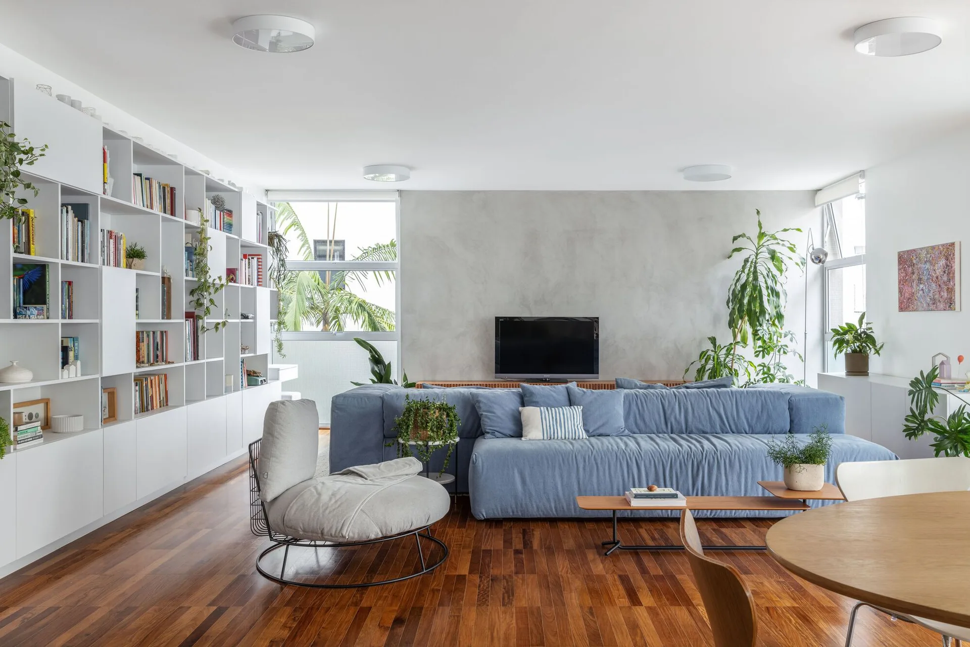 A spacious living room featuring a blue sectional sofa, a concrete accent wall, warm wood flooring, and a large white built-in bookshelf.