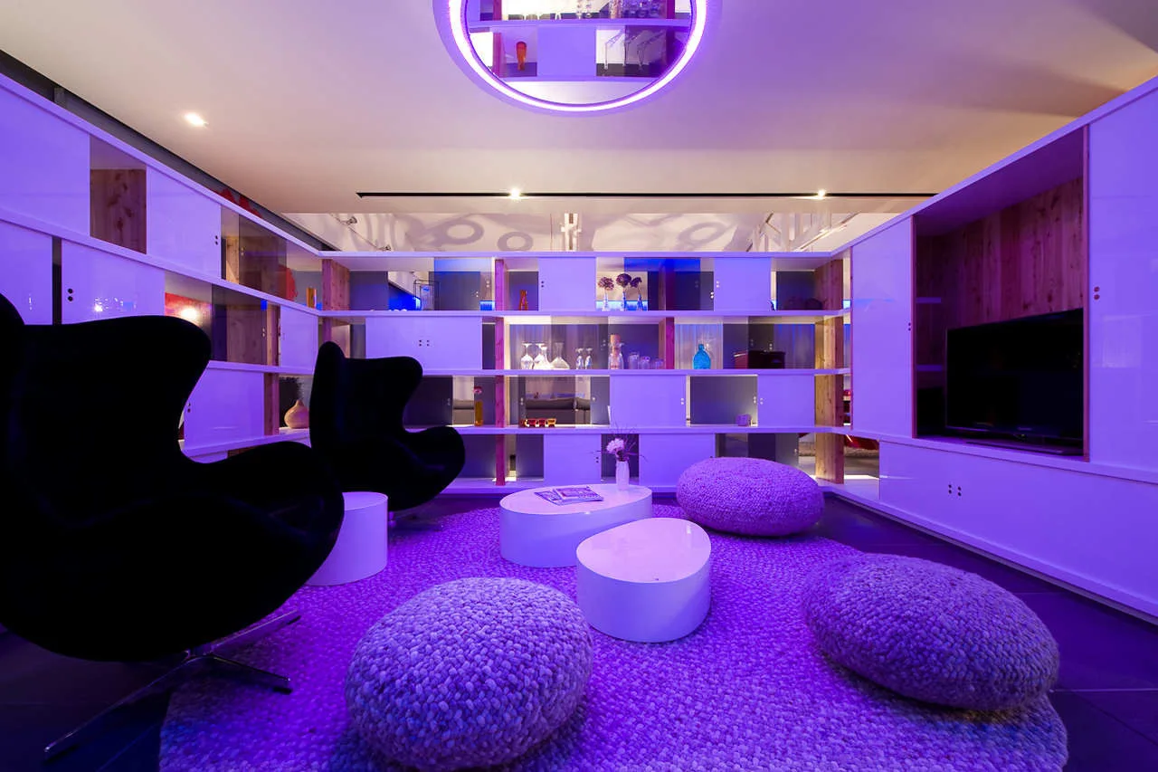A contemporary lounge featuring black egg chairs, textured knit floor cushions, and a custom shelving unit illuminated by purple ambient lighting.