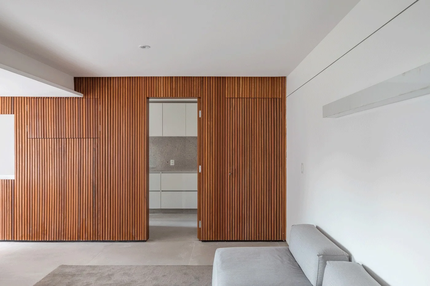 A modern living space features a vertical wood slatted wall, a grey sofa, and an open doorway leading to a white kitchen.