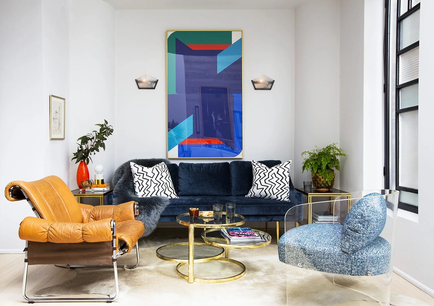 A blue velvet sofa is flanked by a cognac leather chair and a clear acrylic armchair around nested gold coffee tables on a light cowhide rug.
