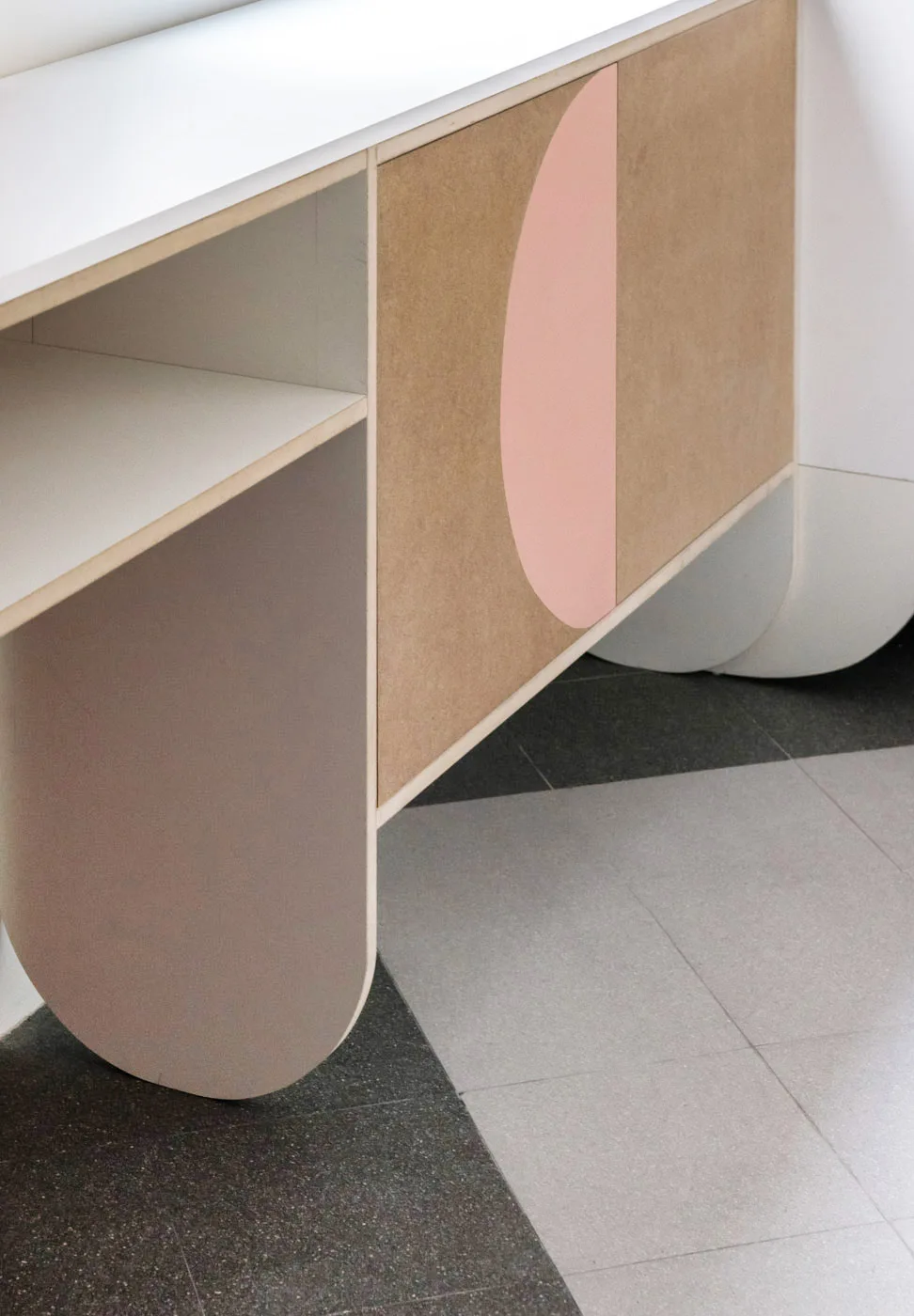 A modern wood cabinet features curved support legs, open shelving, and a flat wood-grain door decorated with a painted pink semicircle on a tiled floor.