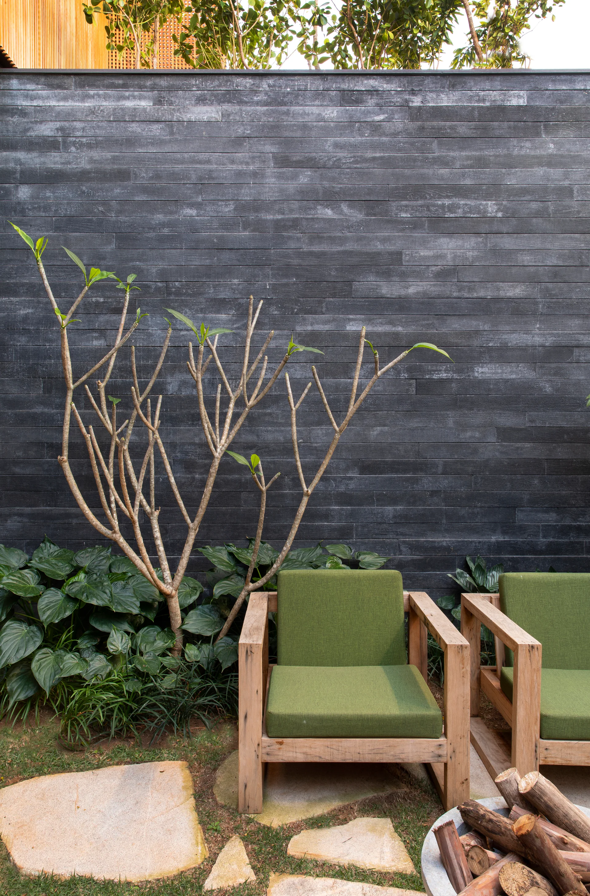 Two wooden armchairs with green cushions sit on a grassy patio with irregular stone pavers in front of a dark charcoal stone wall.