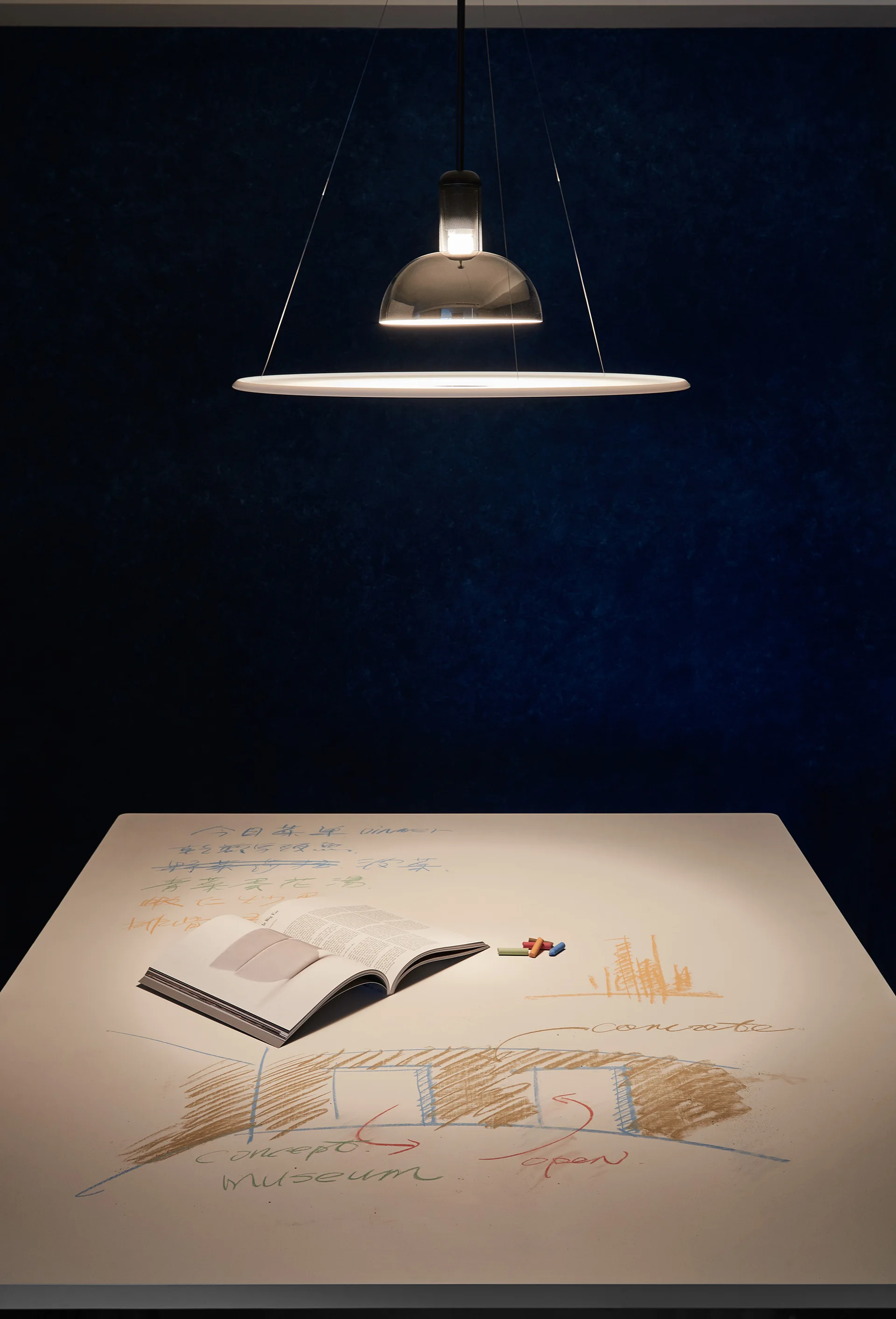 A modern ring pendant light illuminates a white work table covered in sketches and an open book against a textured dark blue wall.