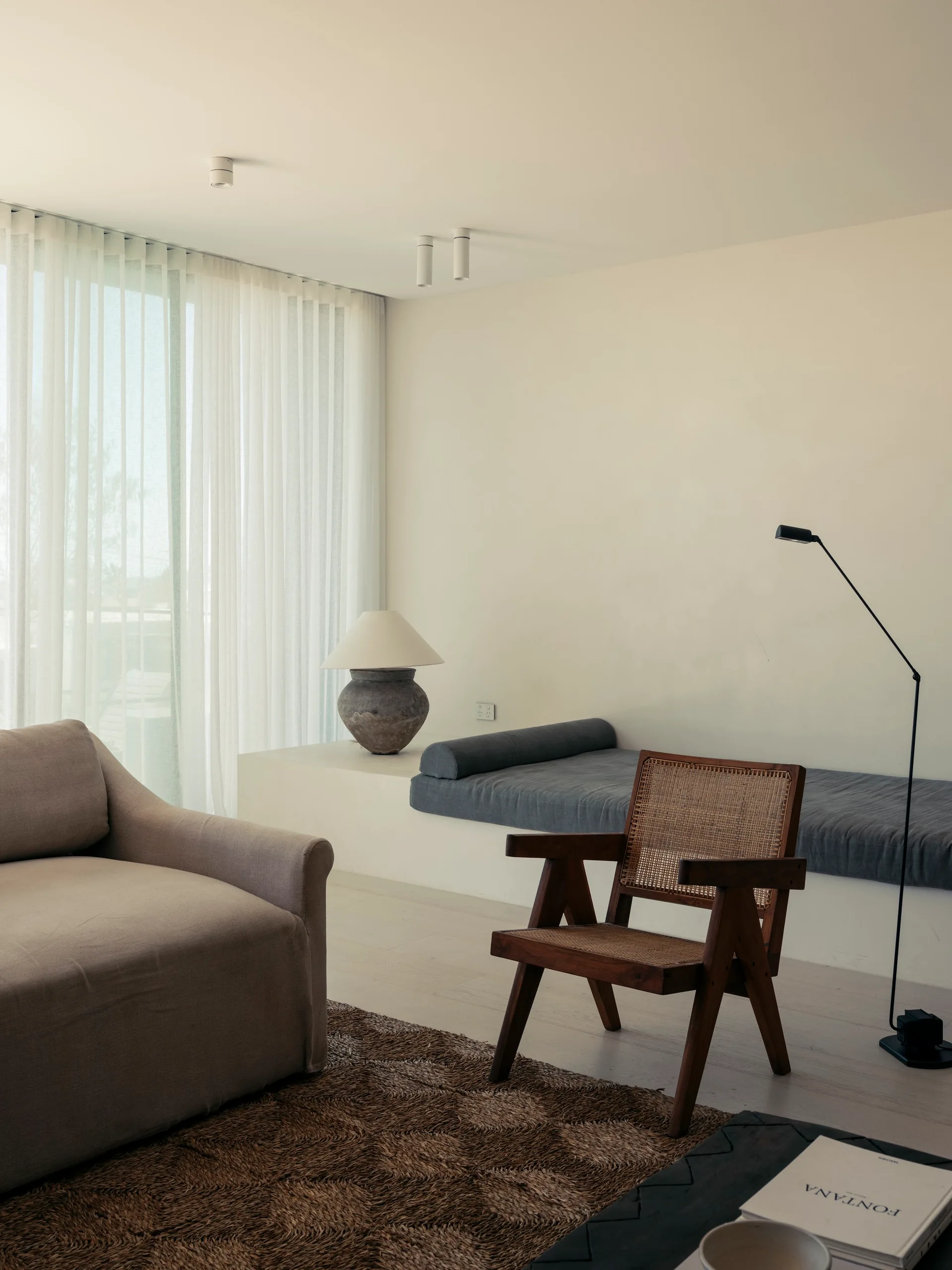 A minimalist living room features a wooden cane armchair, a beige sofa, and a low built-in plaster bench under sheer white window curtains.