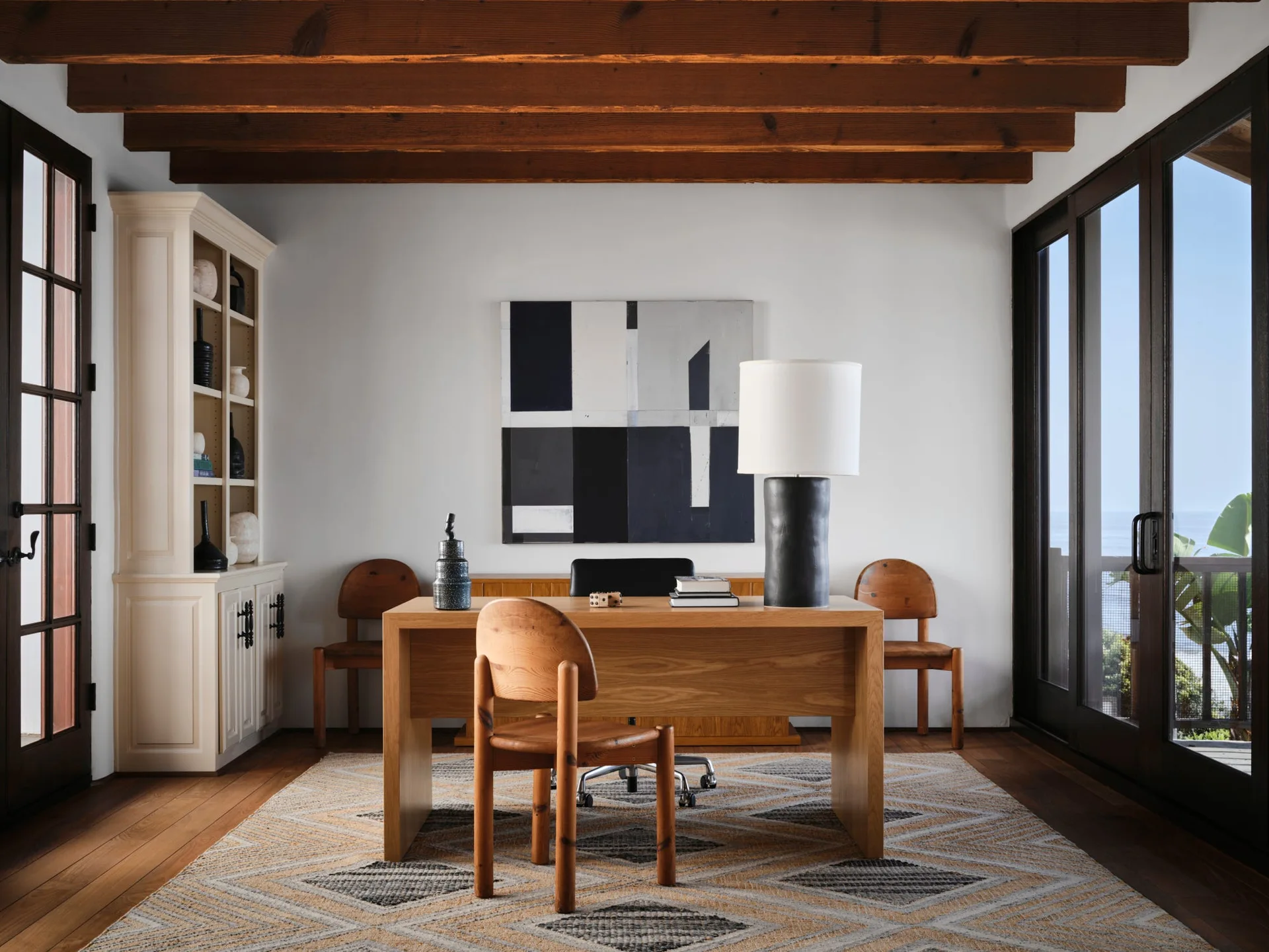 A wooden desk sits on a geometric rug in a home office featuring exposed dark wood ceiling beams, white walls, and a large abstract painting.