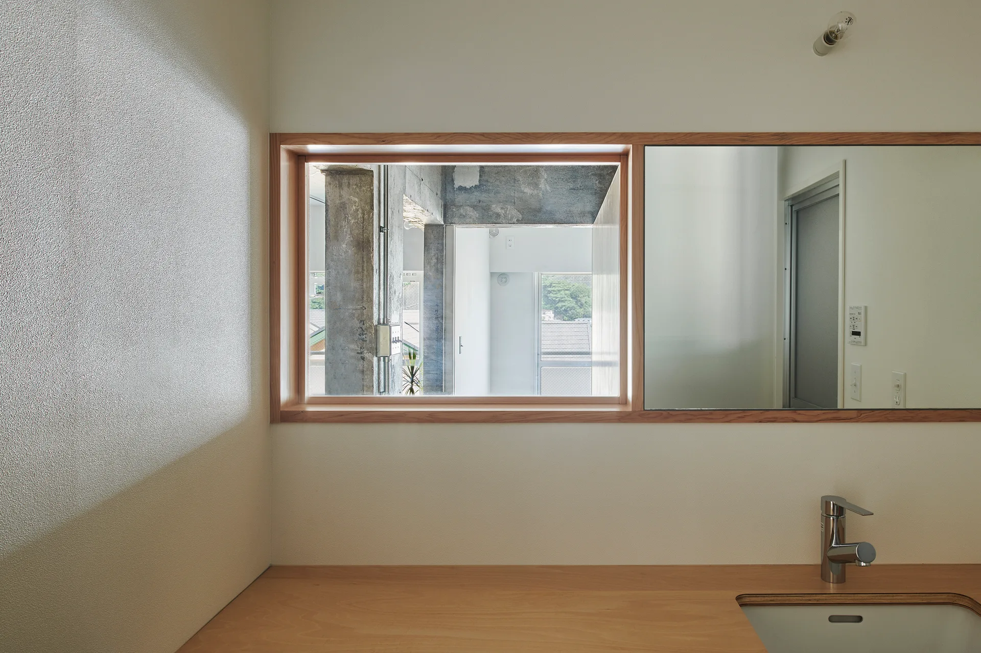 A minimalist bathroom counter with an inset sink, featuring a large wood-framed opening split into an interior window and a matching mirror.