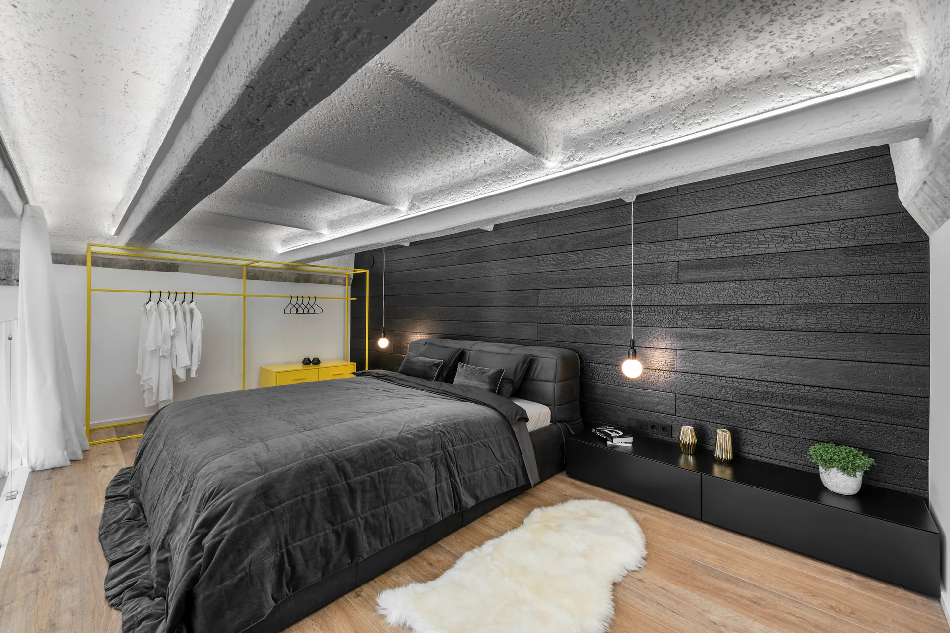 A modern bedroom features a grey bed against a charred wood wall, painted white ceiling beams, and a bright yellow open metal wardrobe.