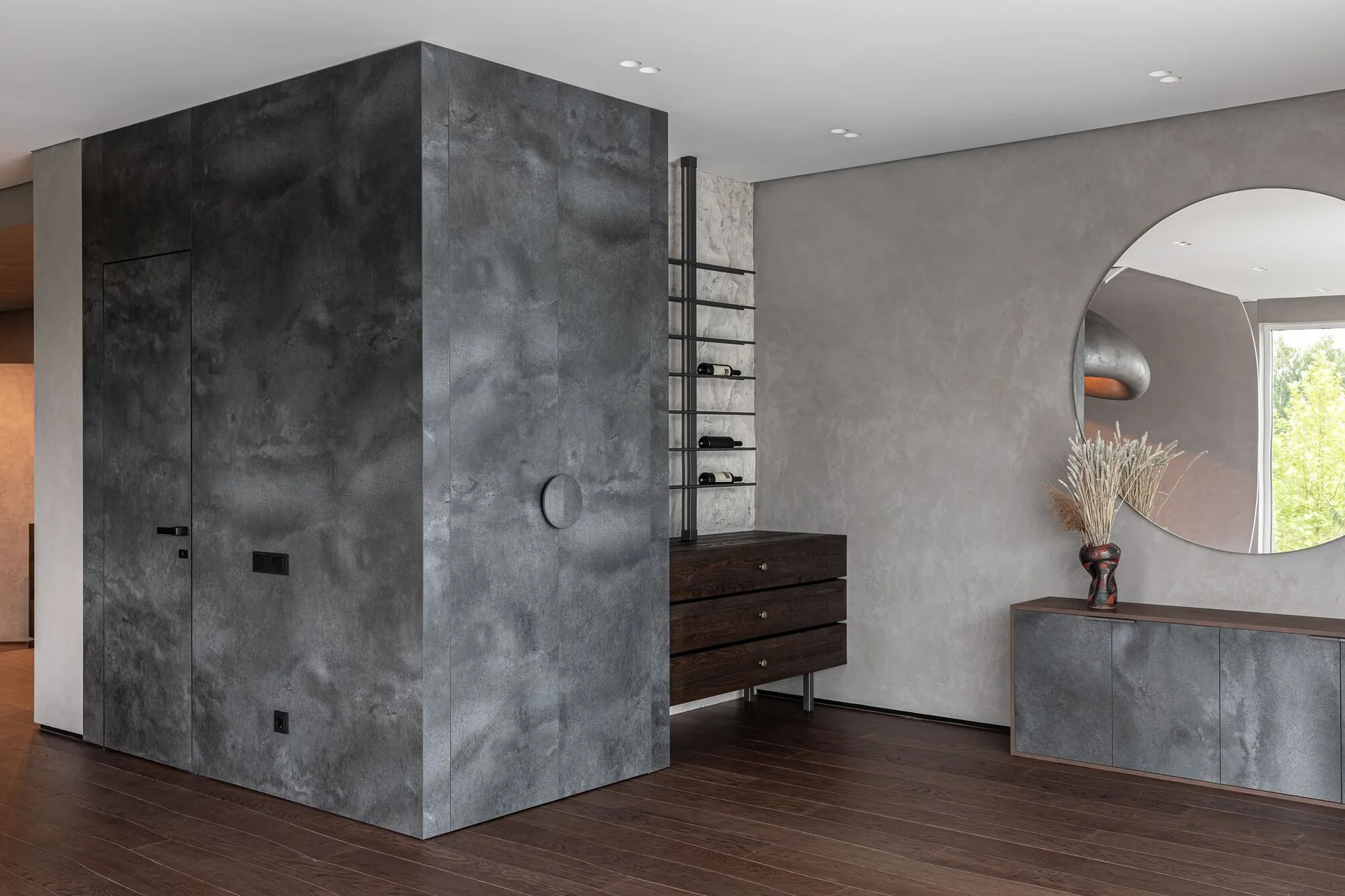 A minimalist room featuring a large charcoal gray storage cabinet, a dark wood dresser, and a round wall mirror on textured plaster.