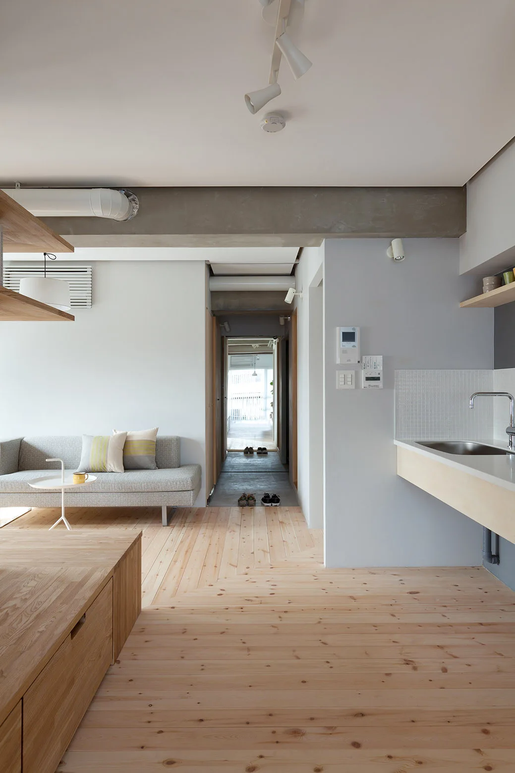 A minimalist open-concept living area features light wood plank flooring, a gray sofa, an exposed concrete ceiling beam, and a kitchen sink counter.