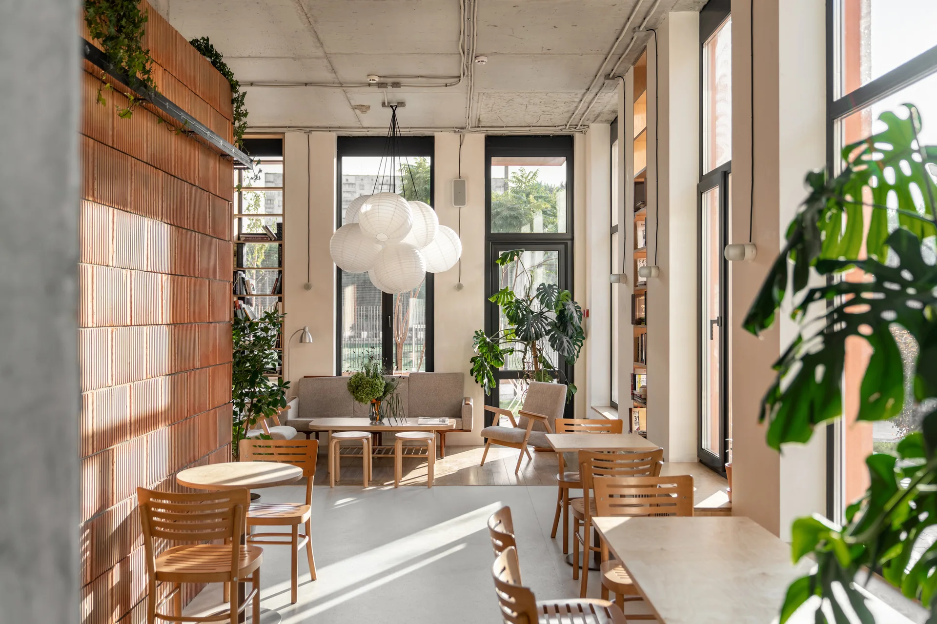 A bright cafe interior featuring a terracotta brick partition wall, wooden tables with chairs, a concrete ceiling, and a cluster of white paper lanterns.
