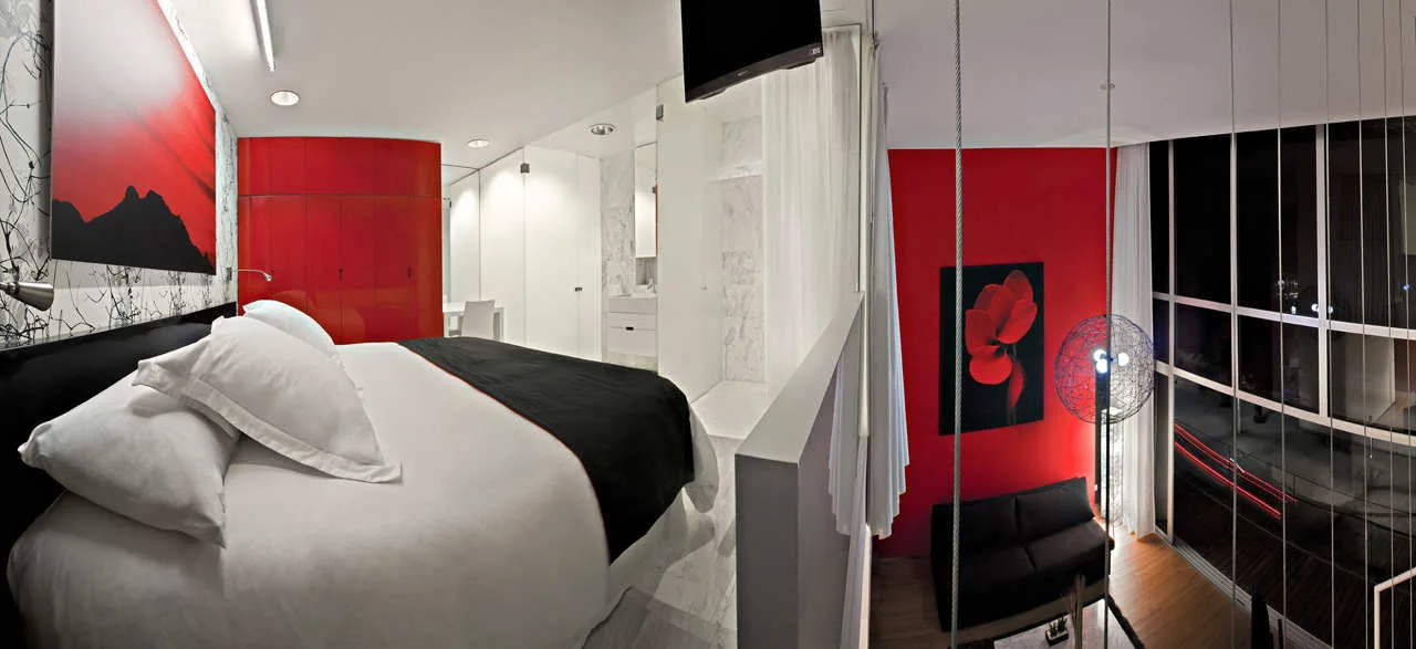A split-level bedroom suite features a large bed with white linens, high-gloss red cabinets, and steps leading down to a living area with a black sofa.