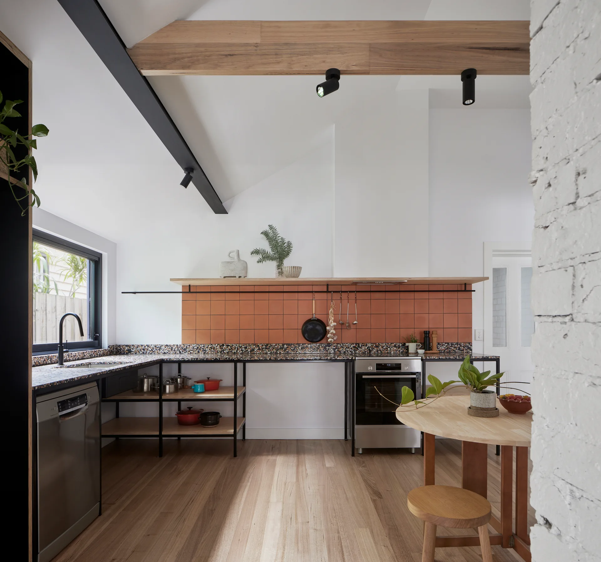 A modern kitchen featuring a terracotta tile backsplash, terrazzo countertops, open lower shelving, and light wood flooring under exposed ceiling beams.
