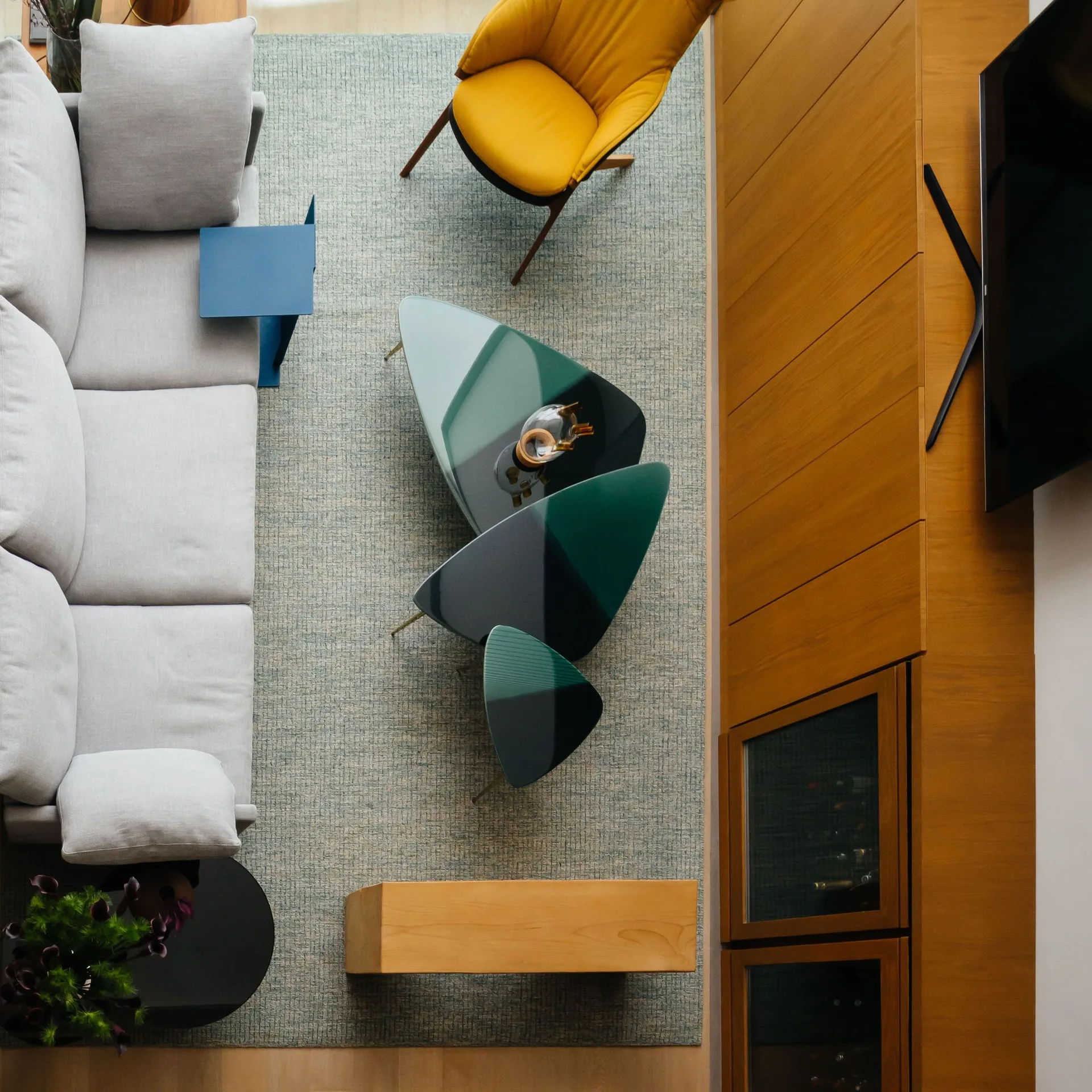 An overhead view of a contemporary living room showing a gray sofa, organic-shaped nesting tables, a mustard yellow armchair, and wood cabinetry.