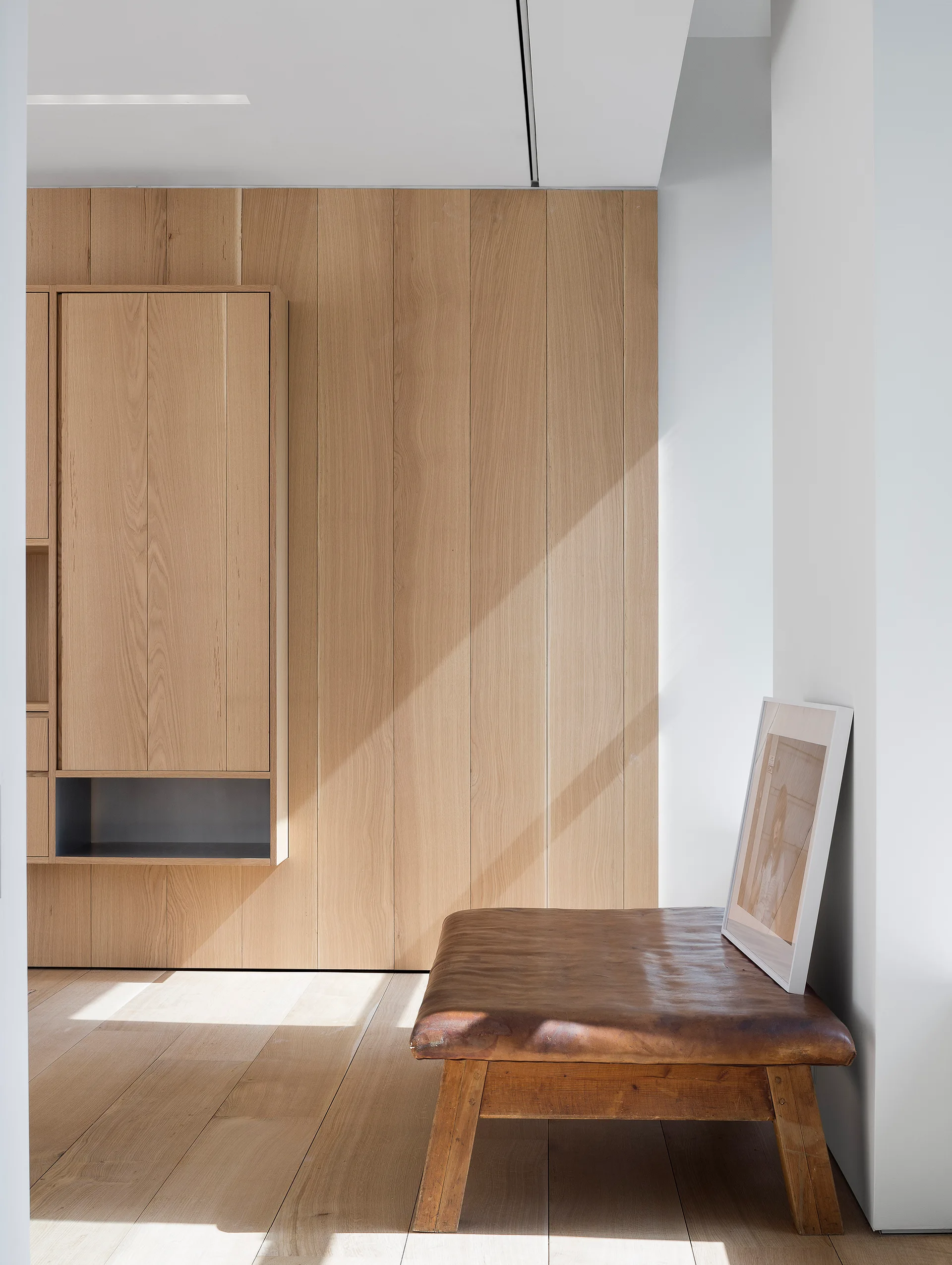 A light oak wood-paneled wall features built-in cabinets next to a worn leather bench with a leaning framed print.