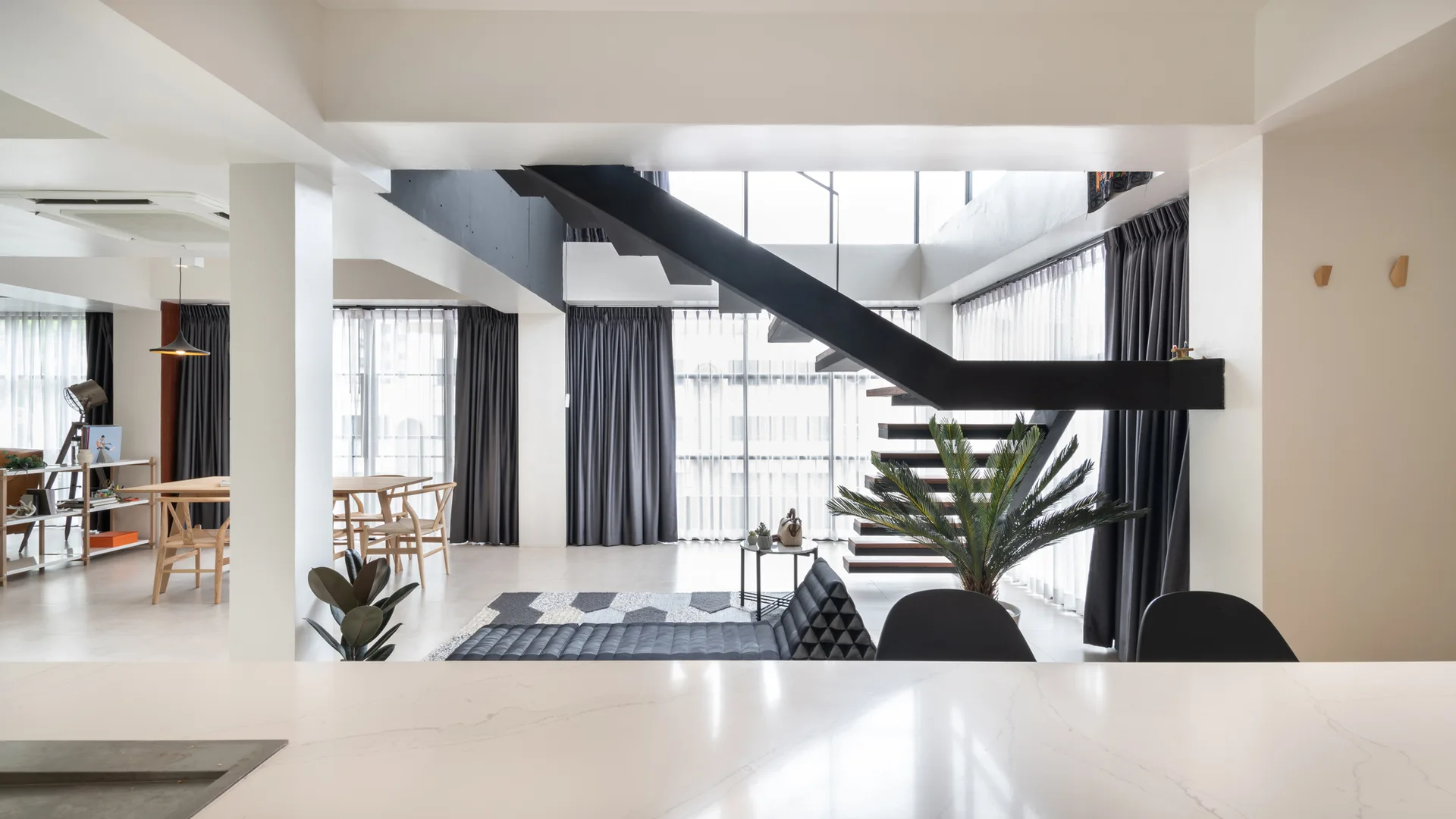 A minimalist open-concept living room featuring a black metal floating staircase, a low-profile grey sofa, and large windows with charcoal drapes.