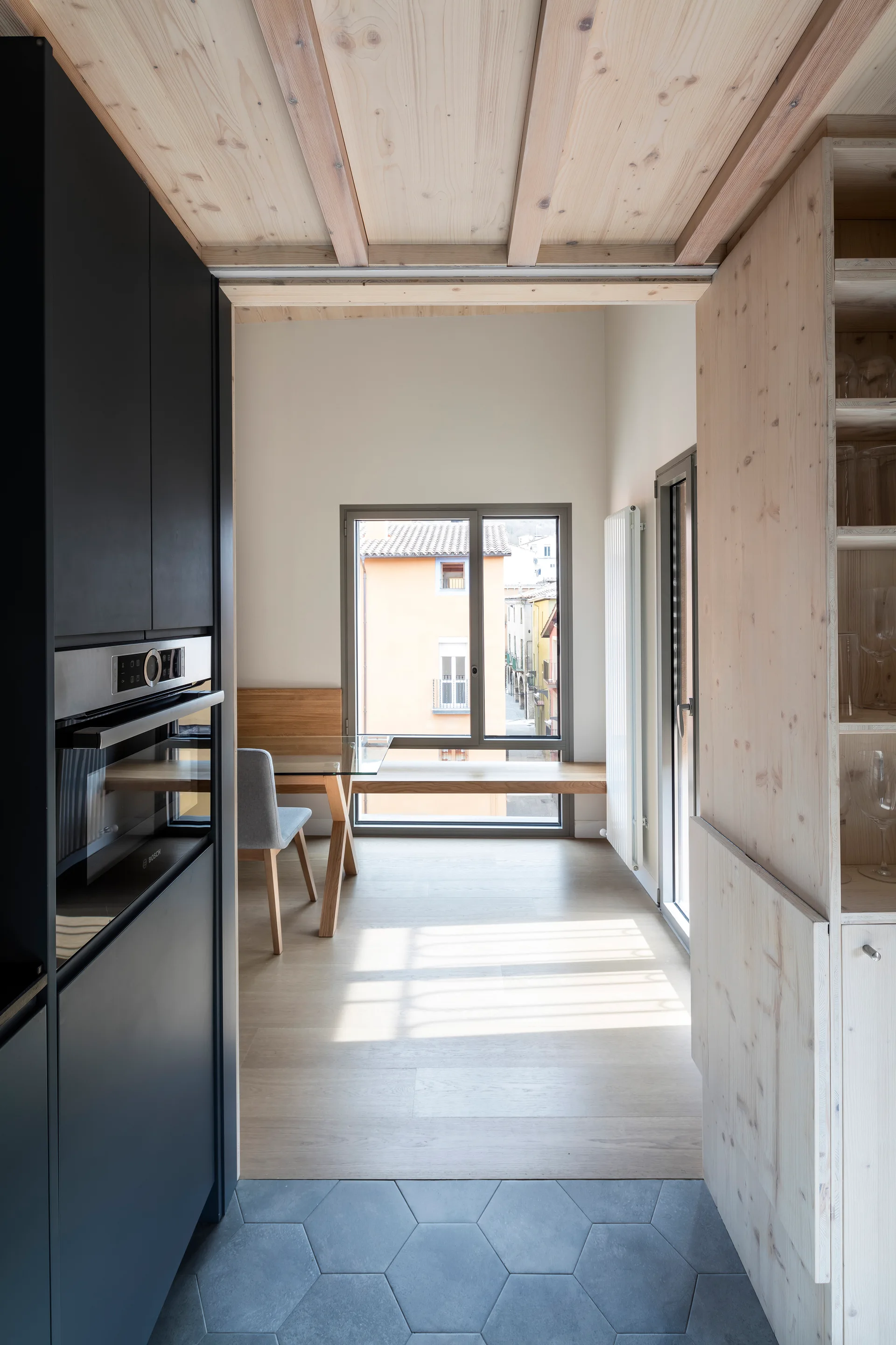 A modern kitchen features matte black cabinets, grey hexagonal floor tiles, a wood-paneled ceiling, and a view into a bright dining area with a window.