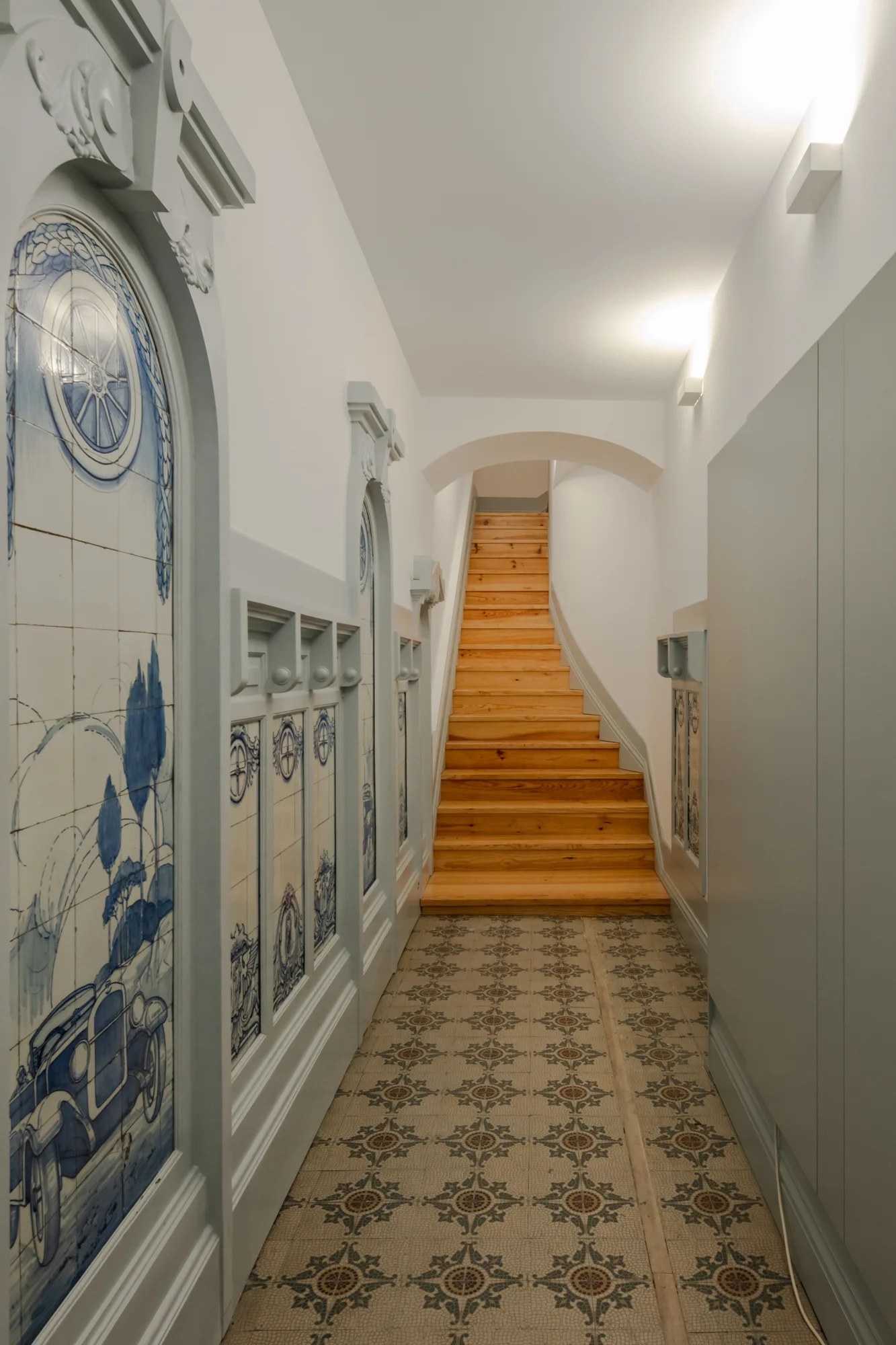 A narrow hallway features blue pictorial wall tiles in light gray frames, patterned floor tiles, and a natural wood staircase under a plaster archway.