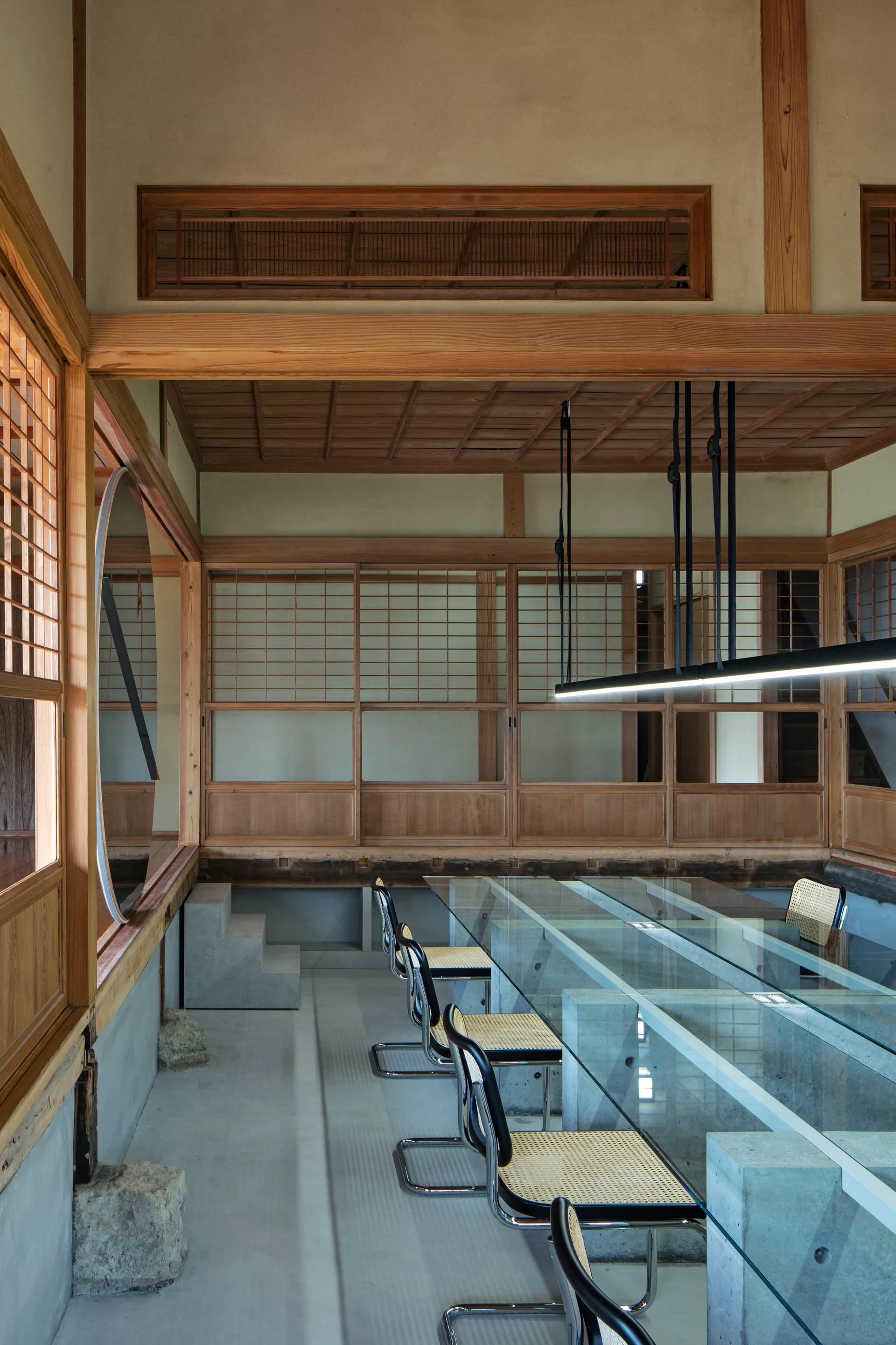 A long glass table with concrete supports is surrounded by modern cane chairs inside a room featuring traditional Japanese wood lattice screens.